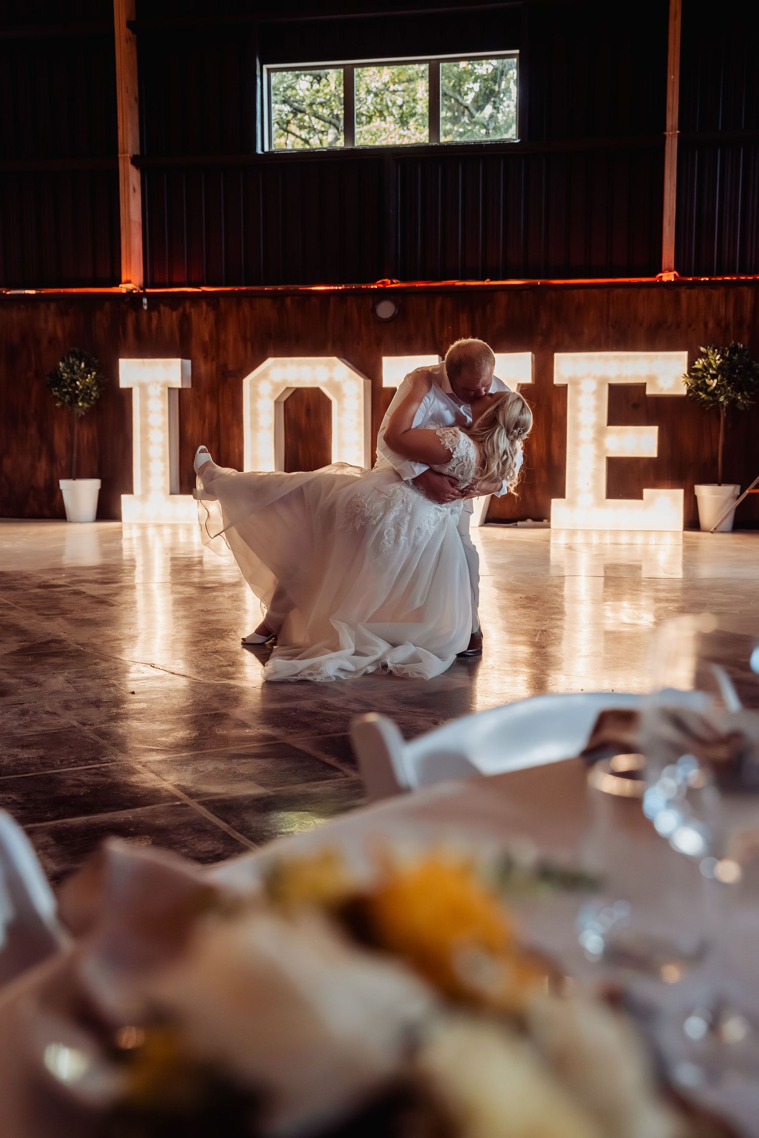 Bride and groom share first dance in front of illuminated LOVE letters at rustic barn wedding reception.