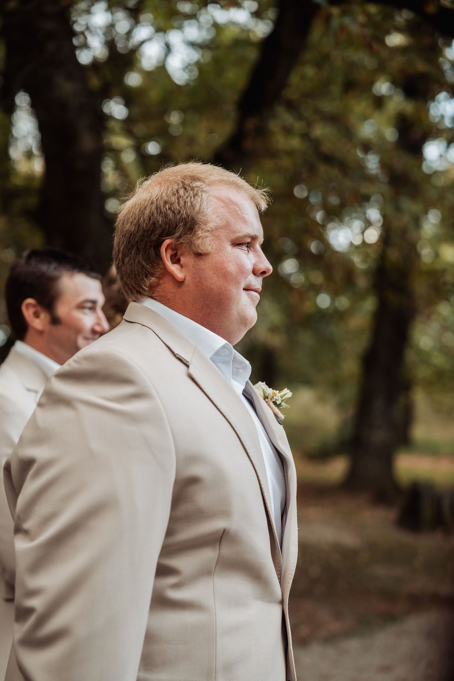 Groom in beige suit with boutonniere standing outdoors among trees at wedding ceremony.