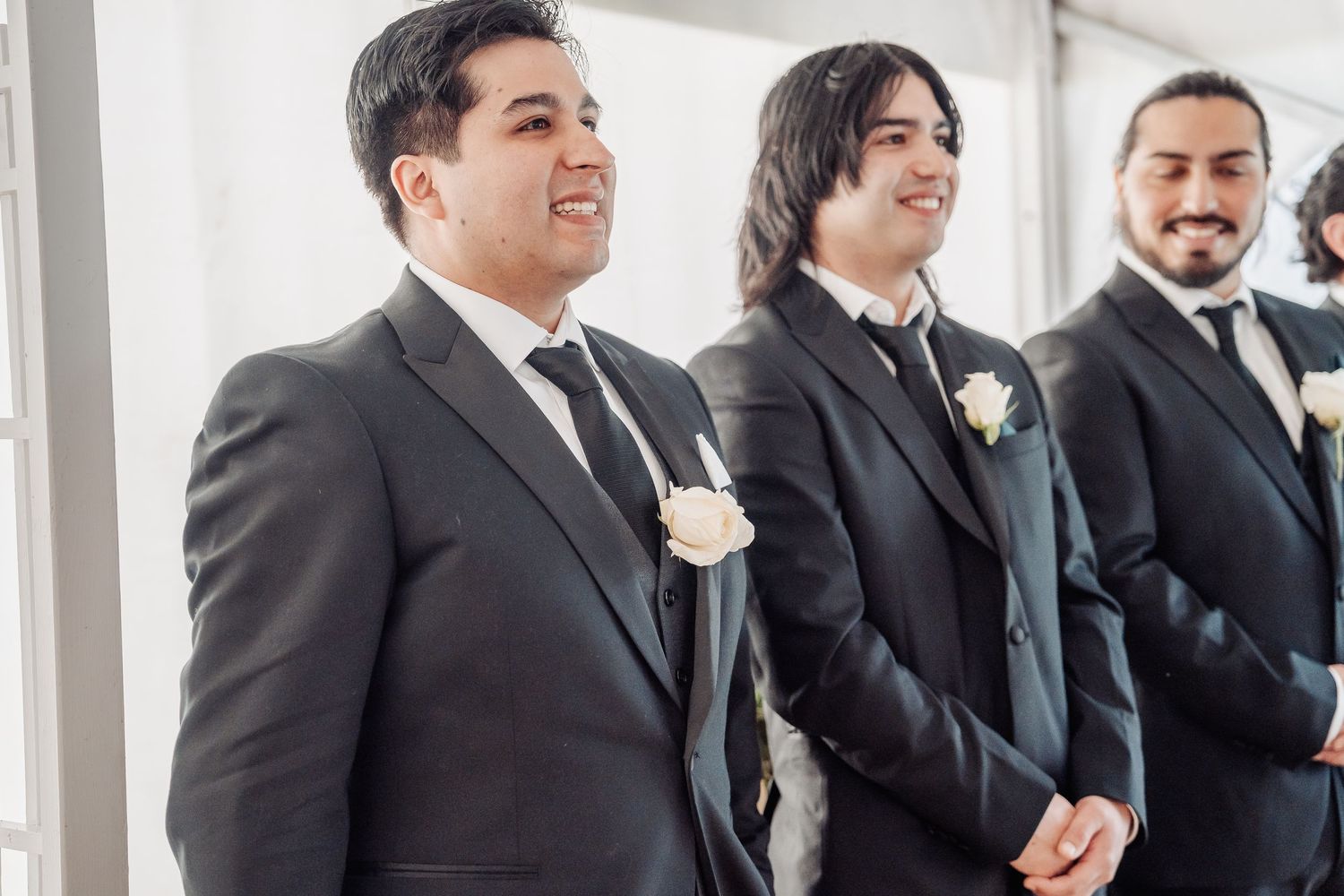 Groomsmen in black formal suits with white boutonniere flowers stand together smiling at a wedding ceremony.