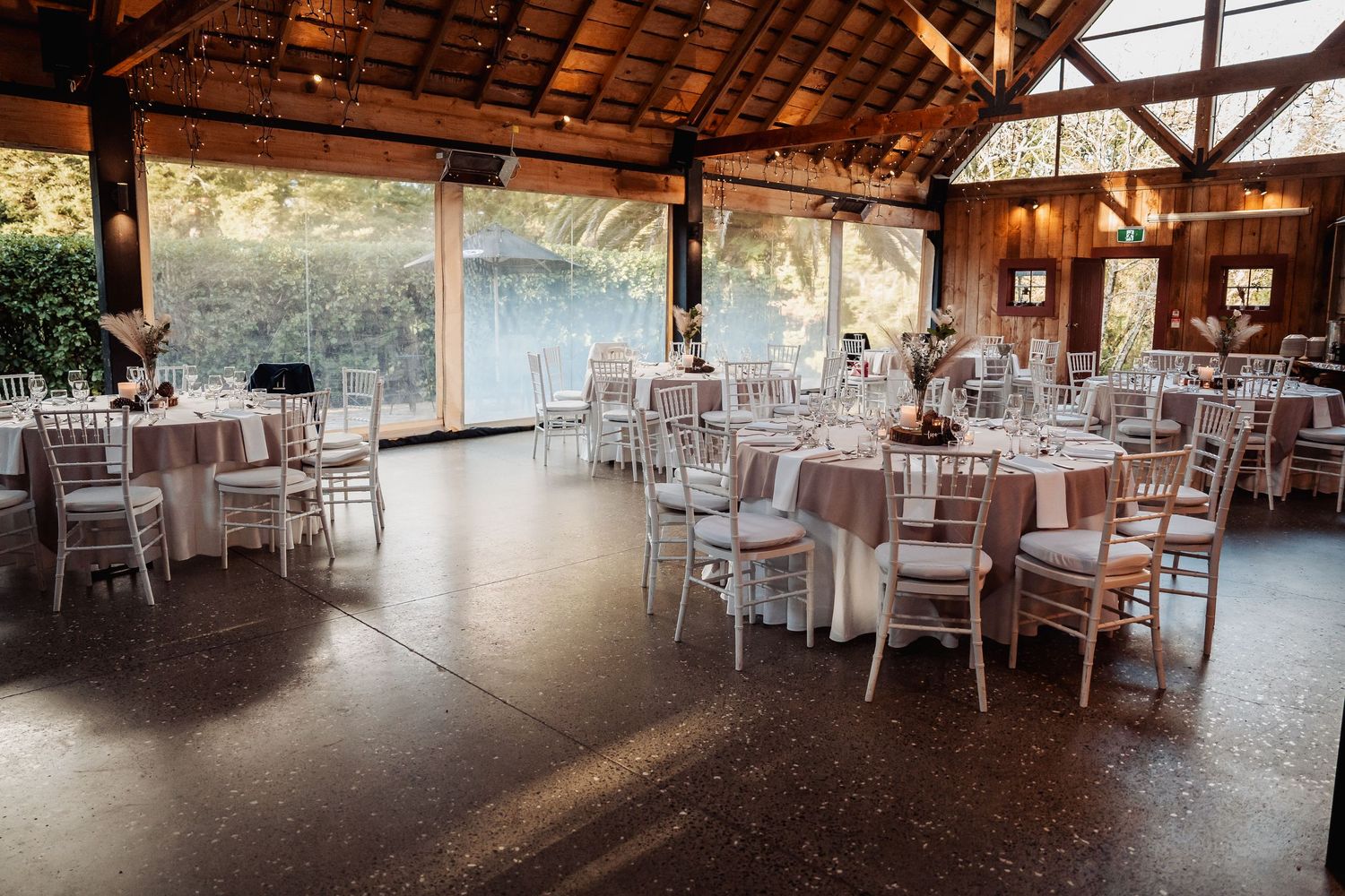 An elegant reception venue with white Chiavari chairs and round tables set up under a rustic wooden beam ceiling.