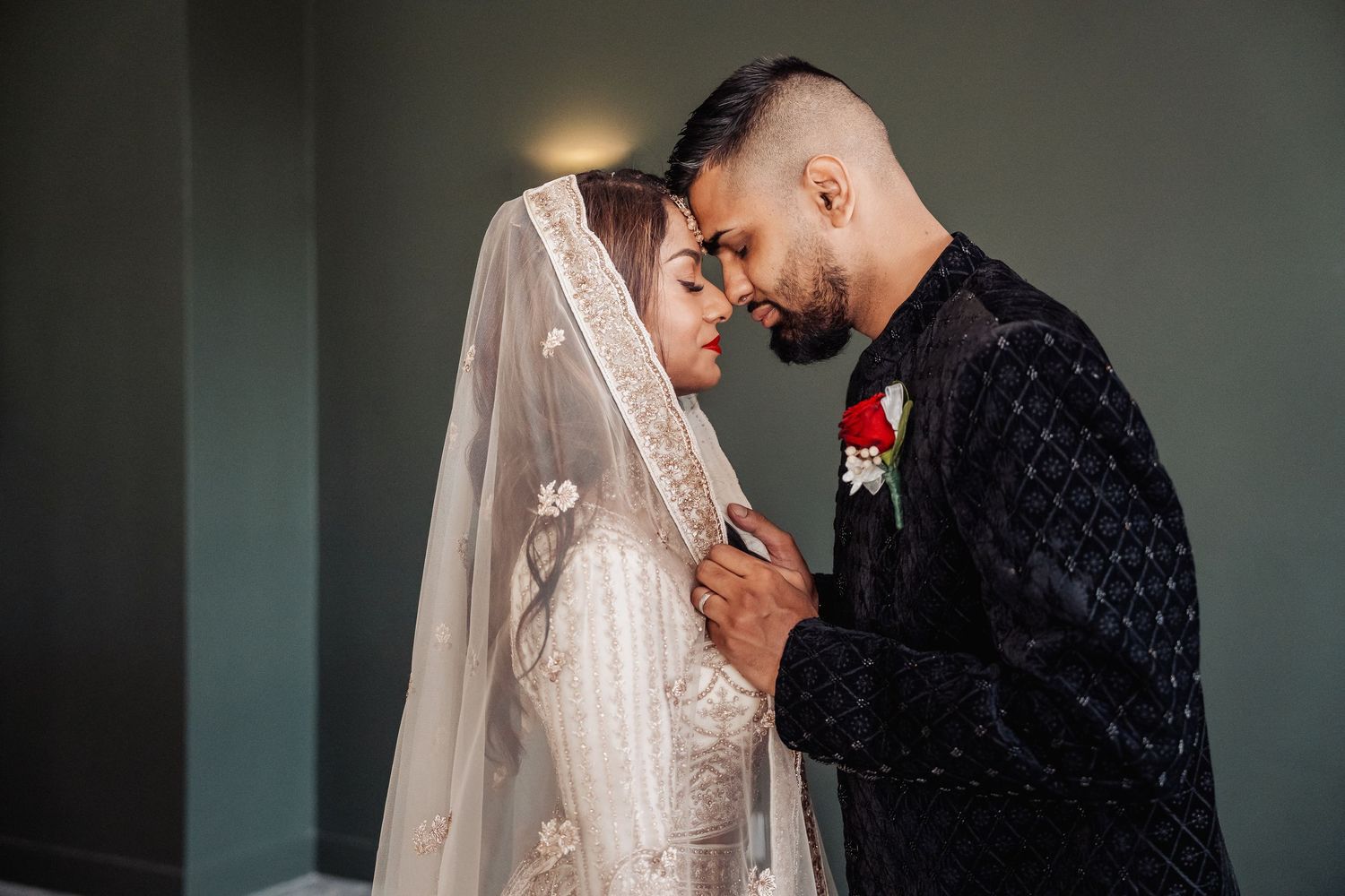 Intimate wedding moment captured against dark green wall with dramatic lighting.
