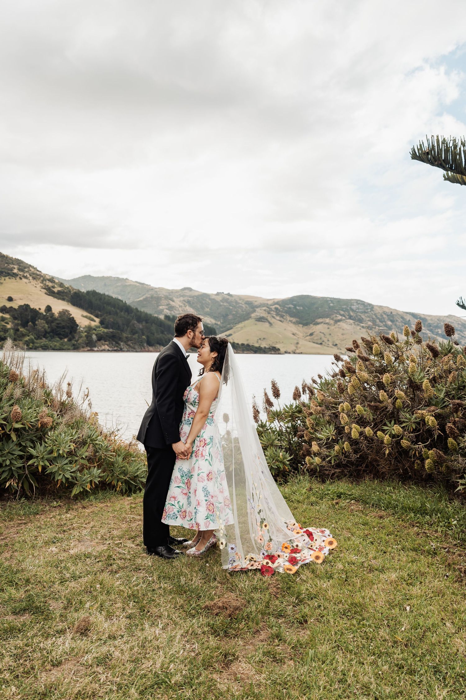 Bride and groom sharing a kiss by a scenic New Zealand lake, bride wearing floral dress with veil.