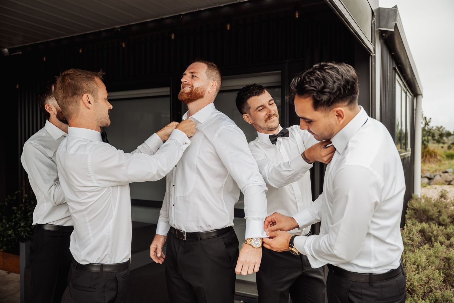 Four groomsmen in white shirts helping each other adjust bow ties outside a modern building.