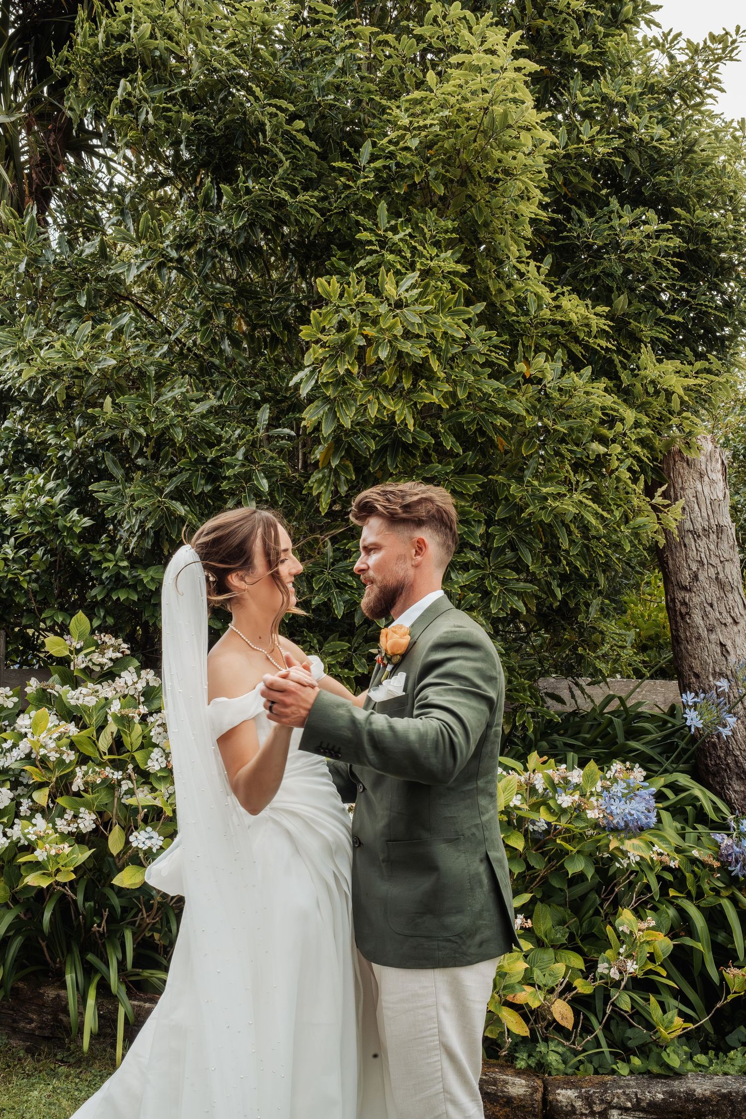 Bride in white gown and groom in green suit share a romantic dance in a flower-filled garden.