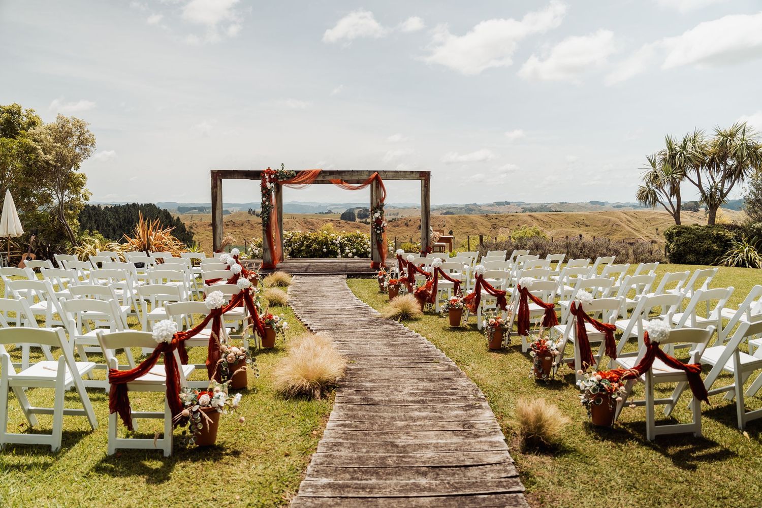 Elegant outdoor wedding ceremony aisle with white chairs and red floral arch overlooking scenic hills.
