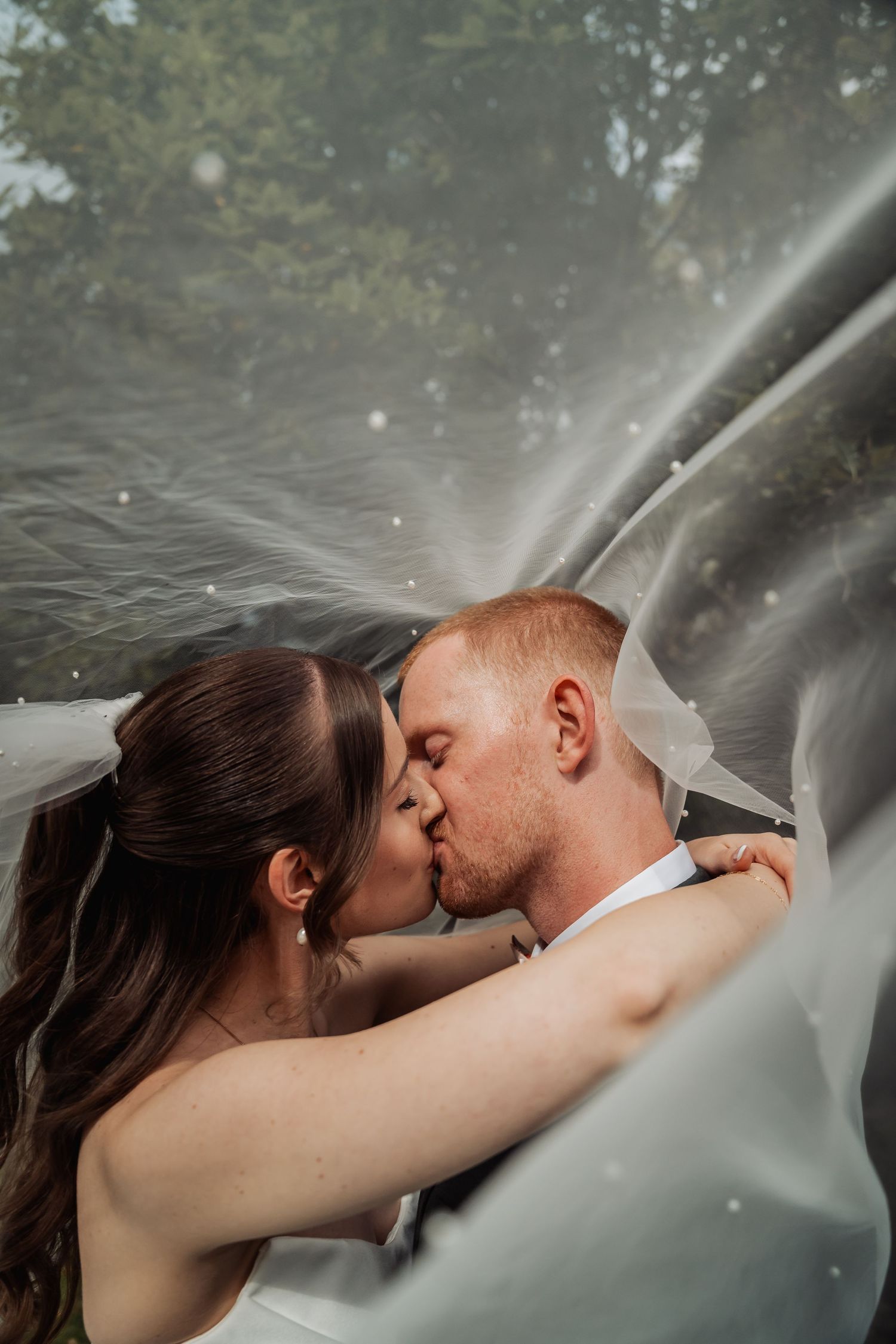 Bride and groom sharing a romantic kiss under a flowing white veil outdoors on their wedding day.