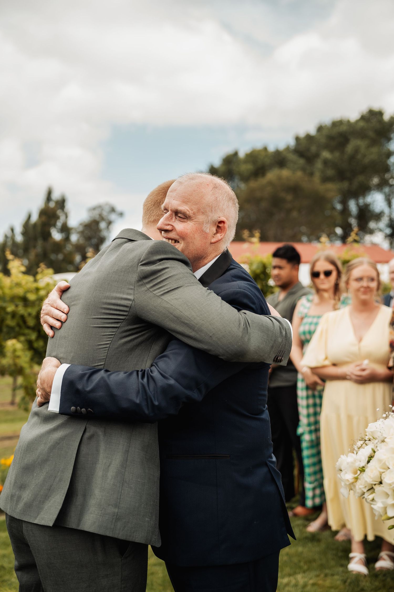 Two men in suits share a warm embrace at an outdoor wedding ceremony.