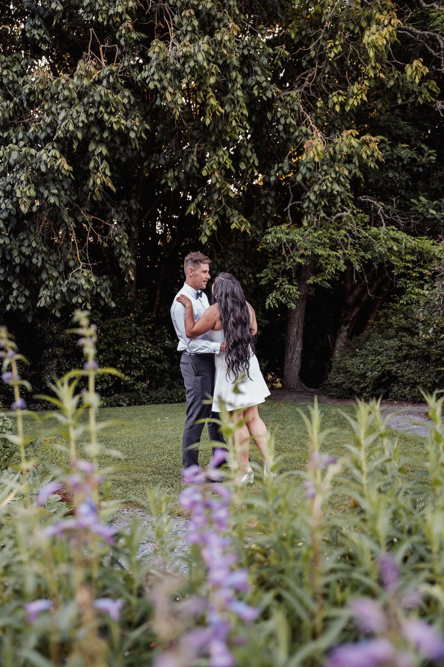 Couple embracing in lush garden with purple wildflowers and green trees in background.