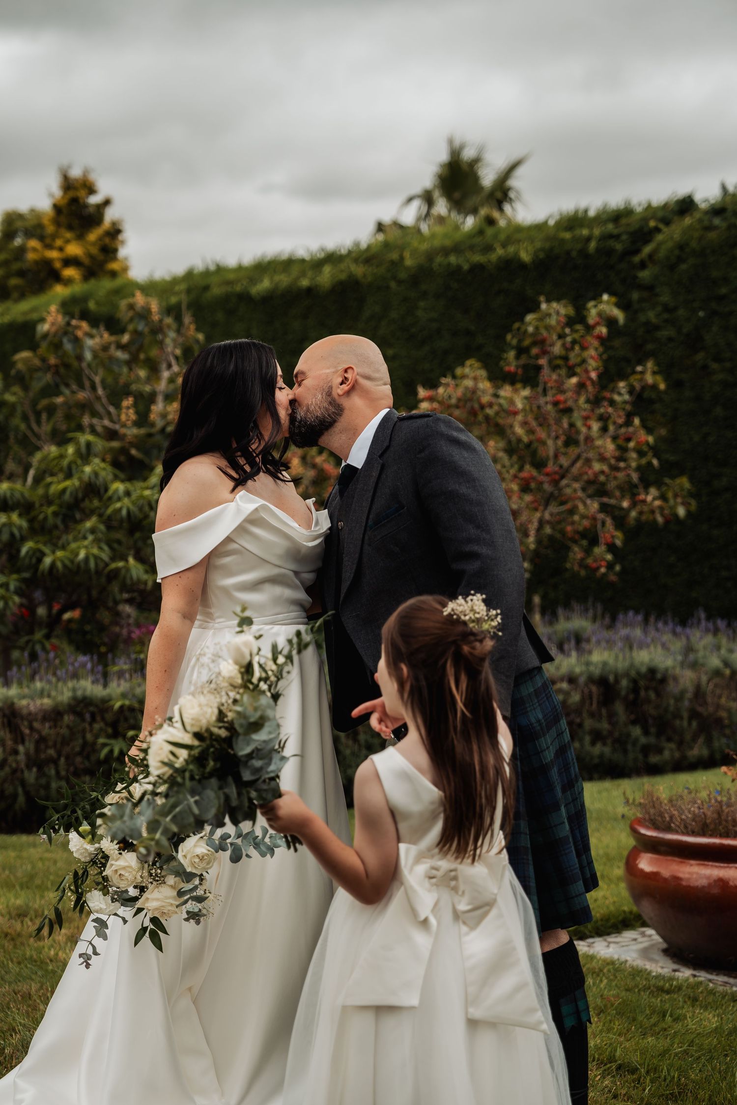Bride and groom sharing a kiss with flower girl in white dress at a garden wedding ceremony.