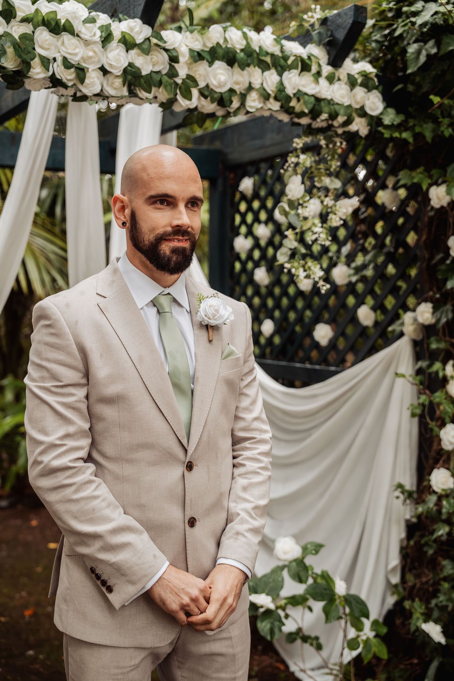 Groom in beige suit with white boutonniere stands under floral arch with white roses at outdoor wedding ceremony.
