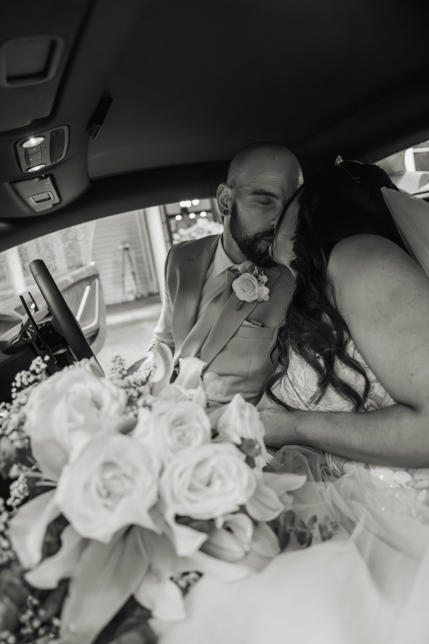Black and white photo of bride and groom kissing in car with bridal bouquet in foreground.
