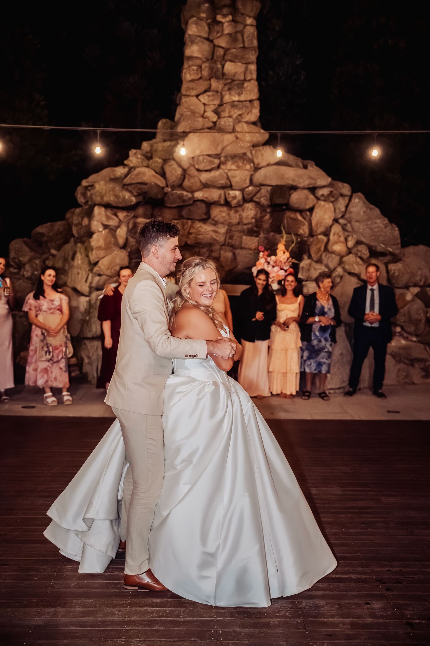 Bride and groom share first dance on outdoor stone patio surrounded by wedding guests at night.