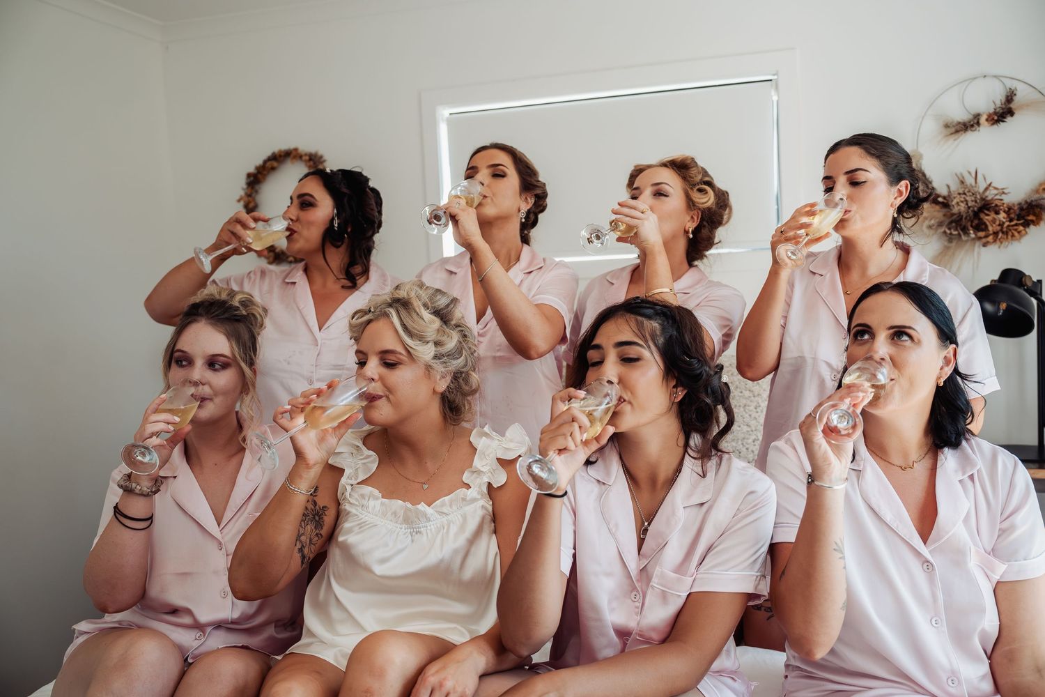 Bride and bridesmaids in pink satin robes drinking champagne together during wedding morning preparations.