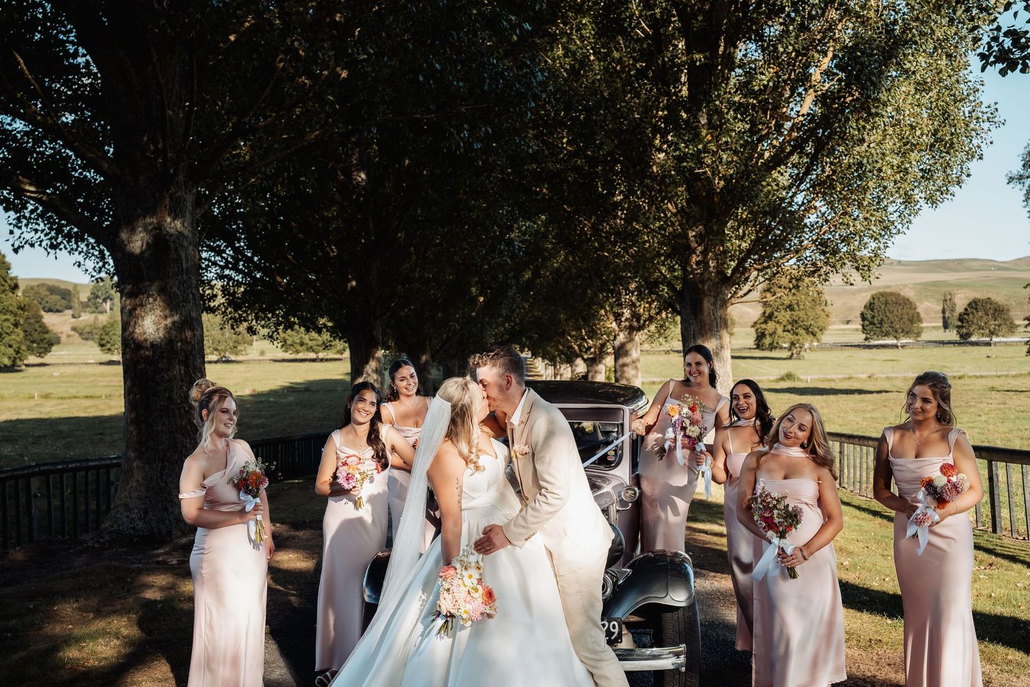 Bride and groom kissing surrounded by bridesmaids near vintage car in scenic landscape.