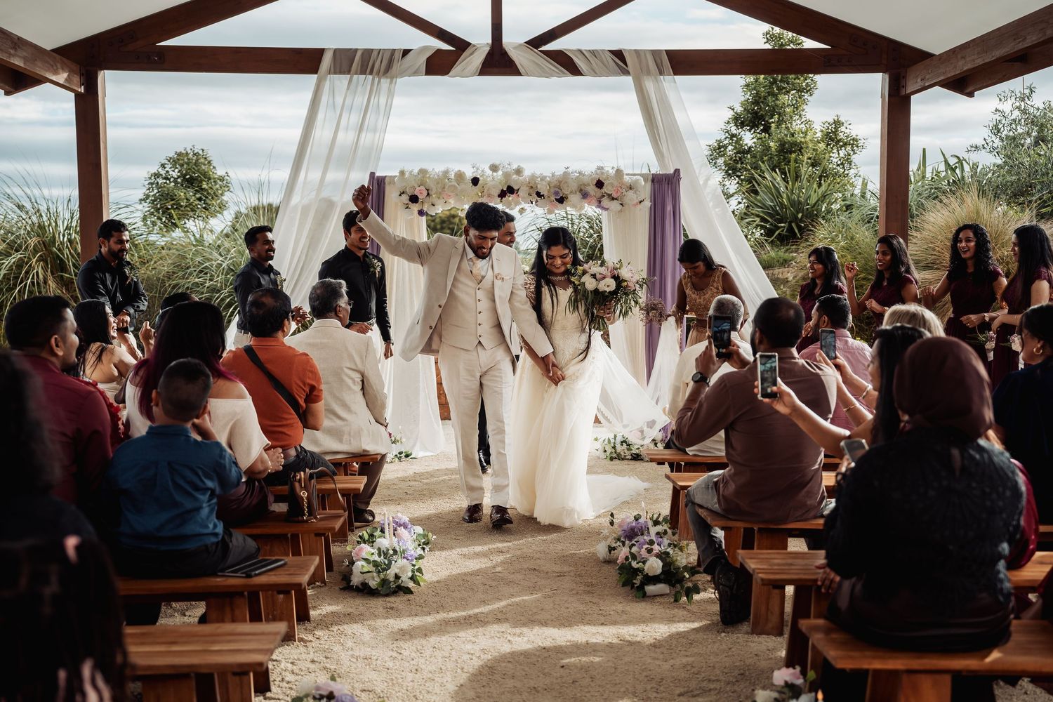 Newlyweds celebrate recessional under floral arch at outdoor wedding with seated guests.