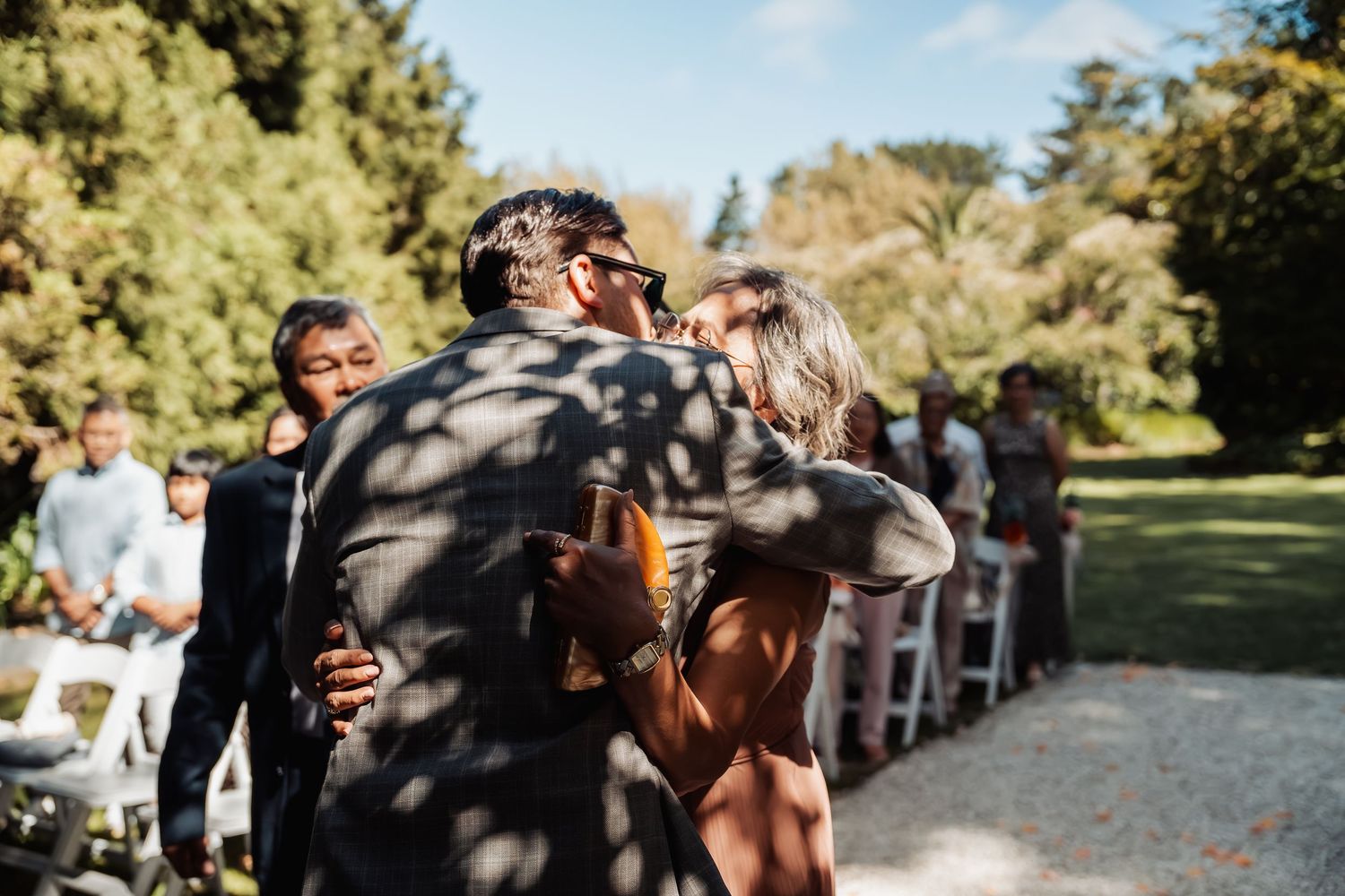 Guests embracing at an outdoor wedding ceremony with white chairs and lush green trees in the background.