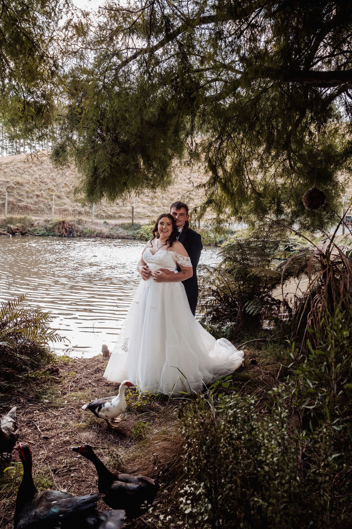 Bride and groom embracing by a tranquil pond surrounded by trees with ducks nearby.