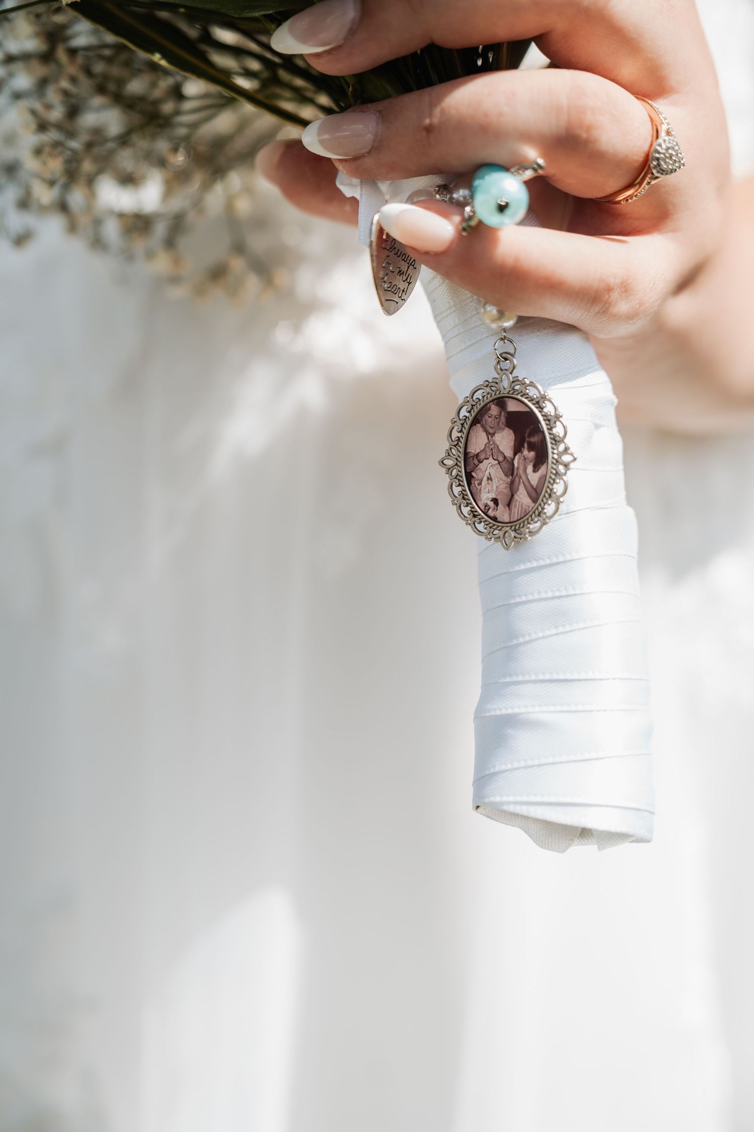 Bride holds bouquet with sentimental oval locket charm and ribbon, wearing turquoise and gold rings.
