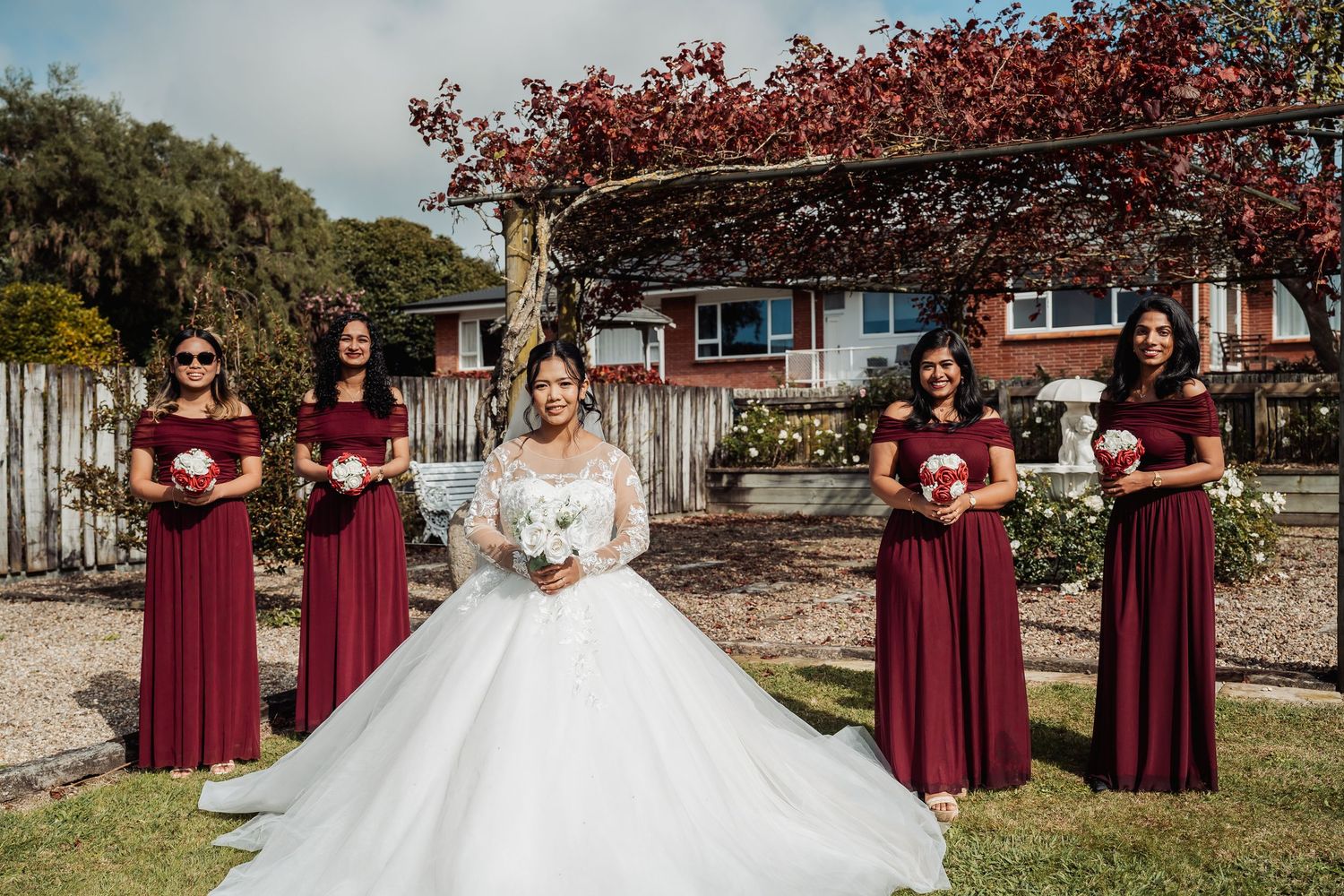 Bride in white ball gown with four bridesmaids in burgundy dresses holding bouquets outdoors.