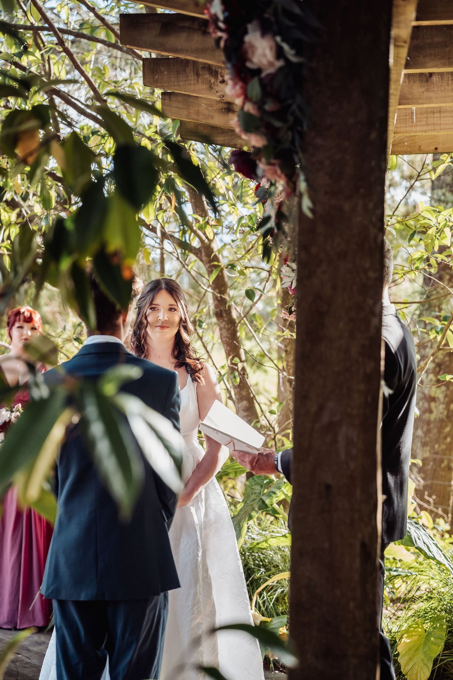 Bride and groom exchange vows under rustic wooden arch decorated with flowers at ceremony.