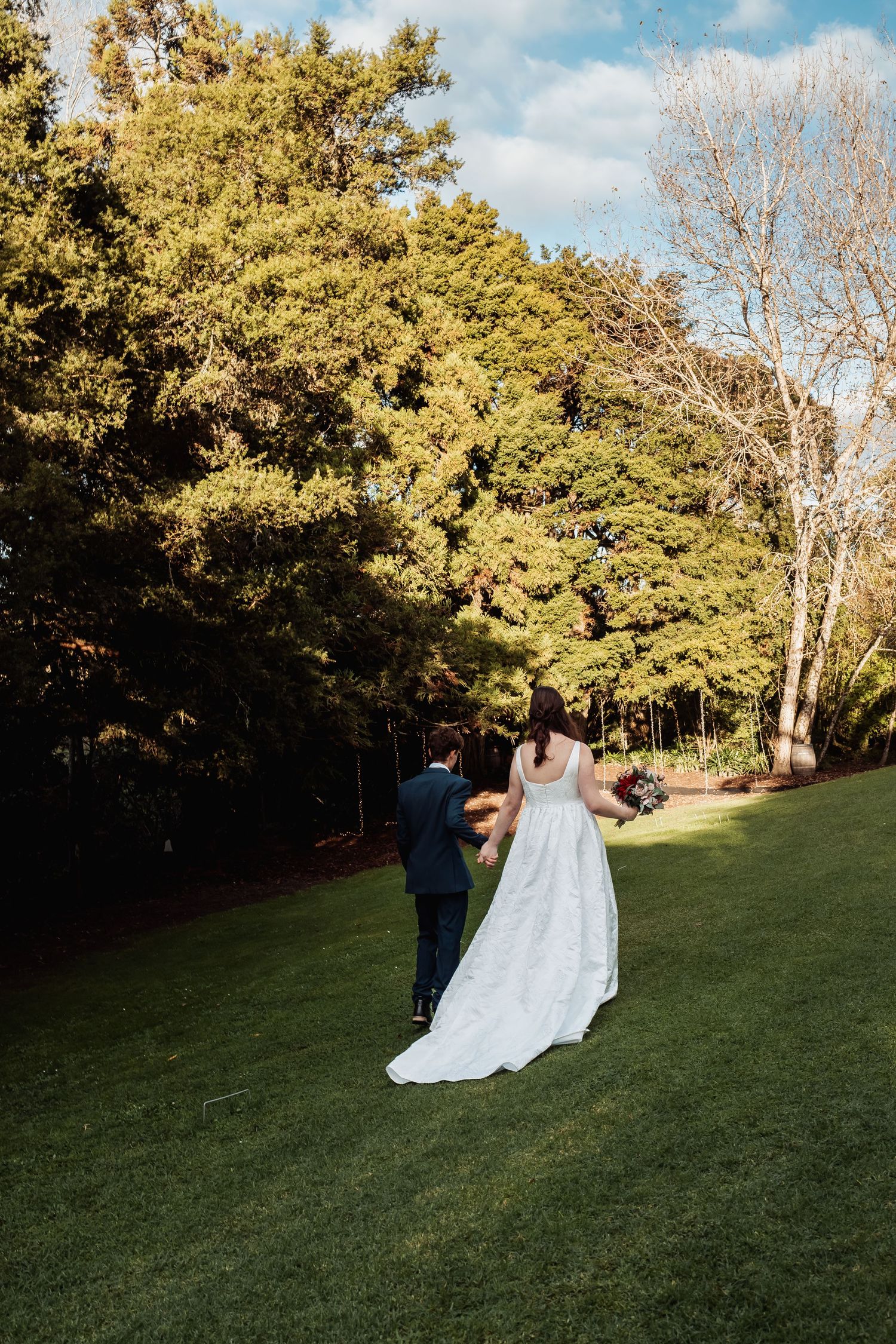 Bride and groom walk hand in hand across green lawn surrounded by trees.