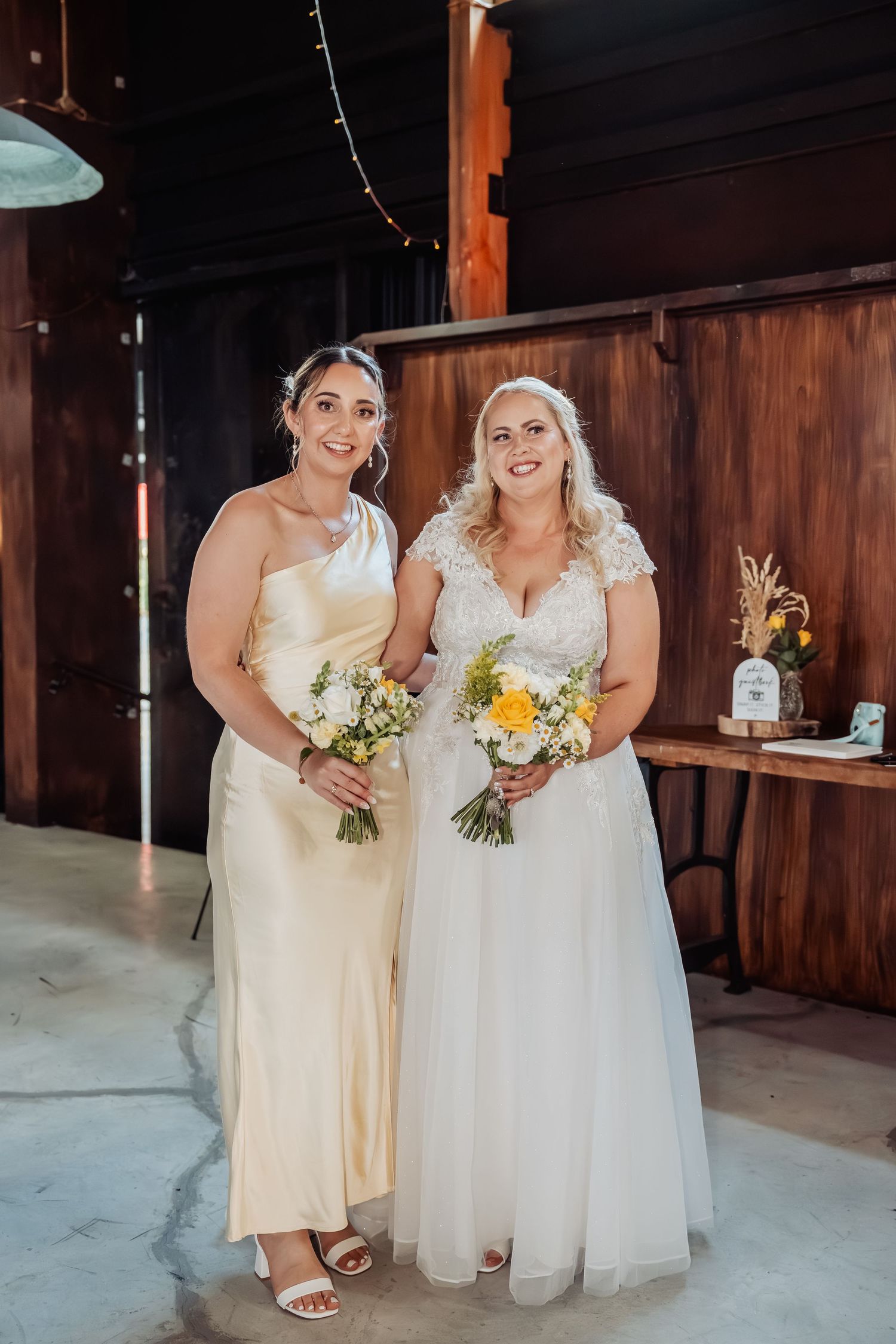 Bride in white lace gown and bridesmaid in yellow satin dress holding bouquets at rustic barn wedding venue.