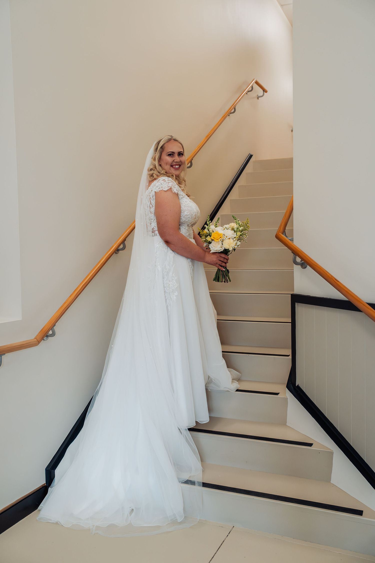 Bride in white lace gown holding bouquet on staircase.