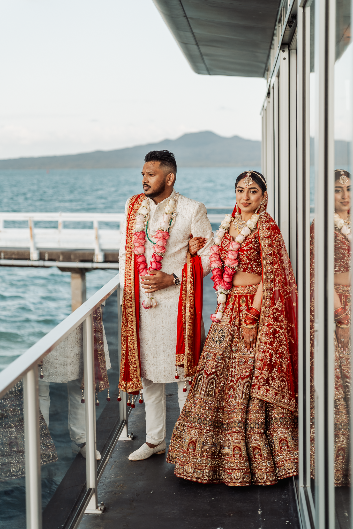Couple in traditional Indian wedding attire posing on oceanfront balcony with mountain backdrop.