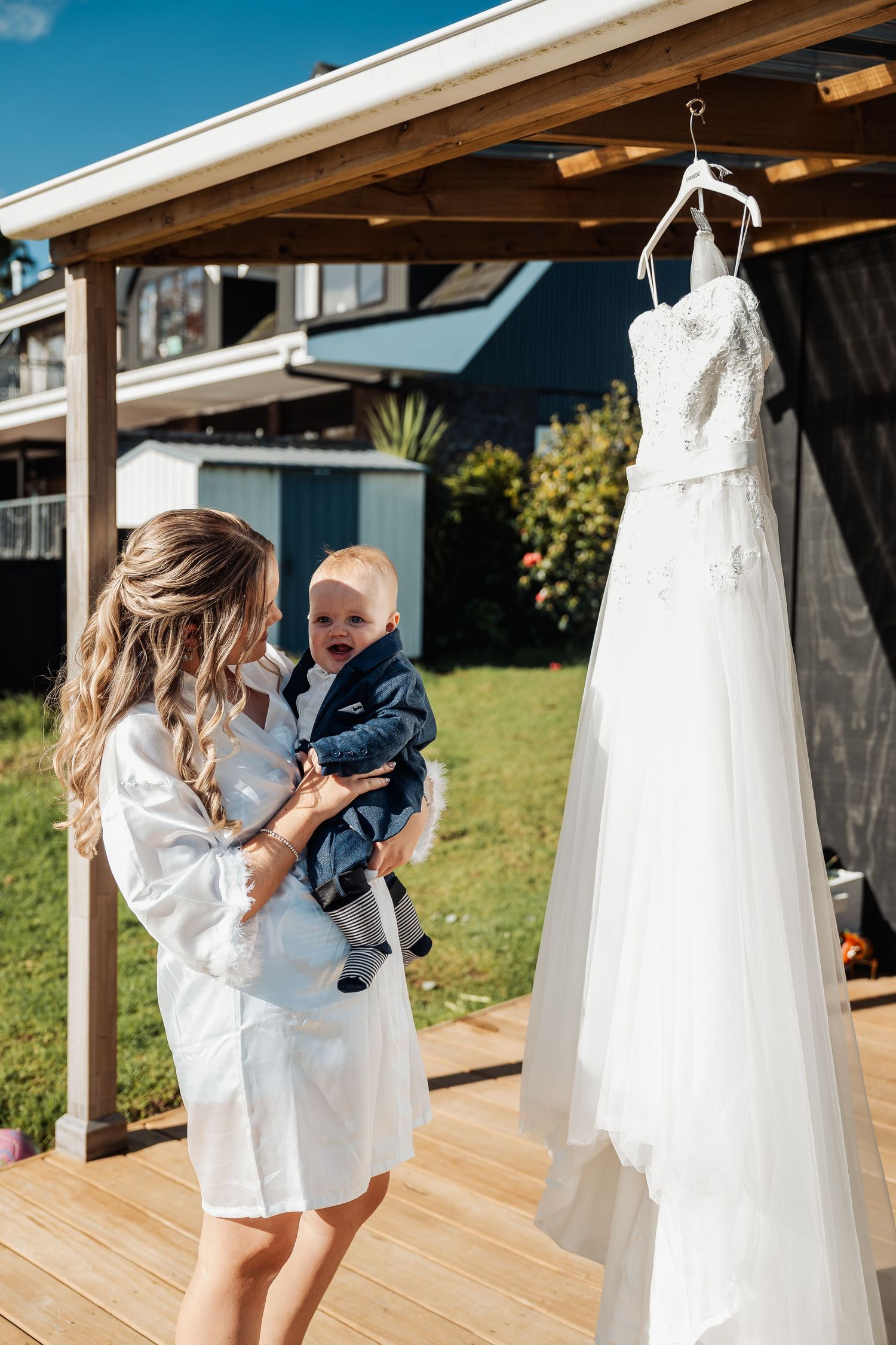 A white wedding dress hangs on display while someone holds a small child on a wooden deck under a covered porch area.