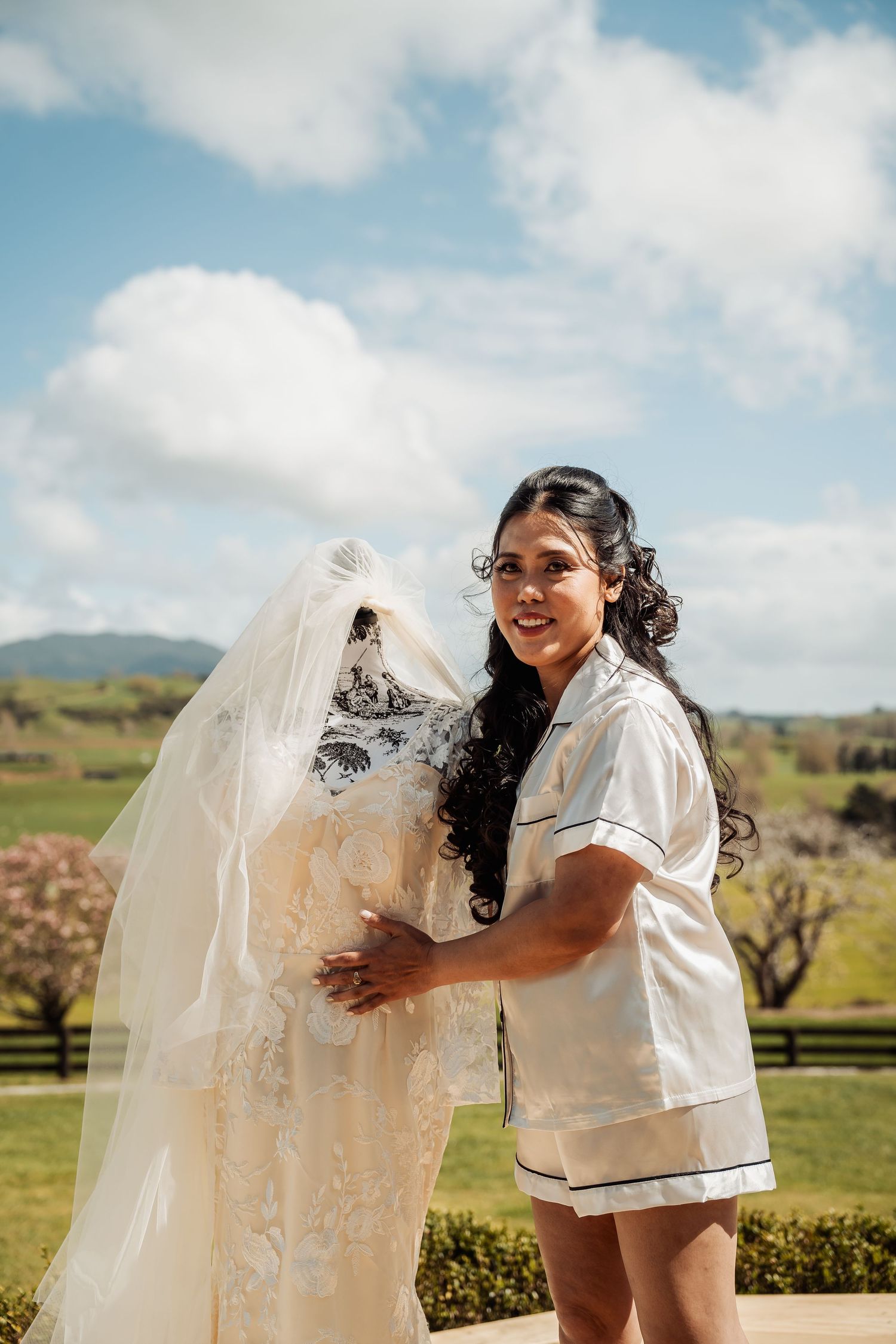 Playful wedding party poses outdoors with flowing veil against scenic countryside backdrop.