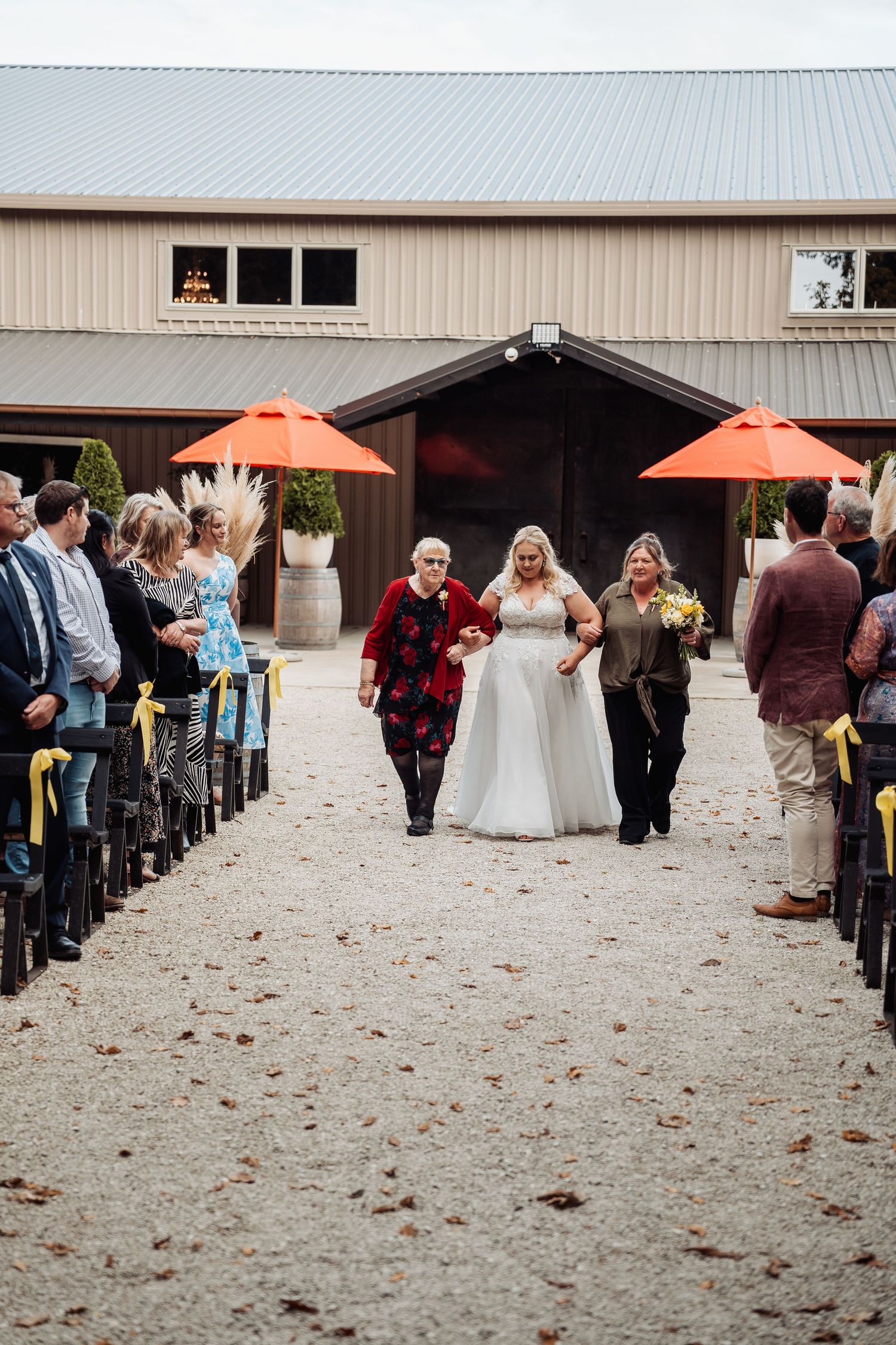 Bride in white gown walking down outdoor aisle with two escorts at barn venue.