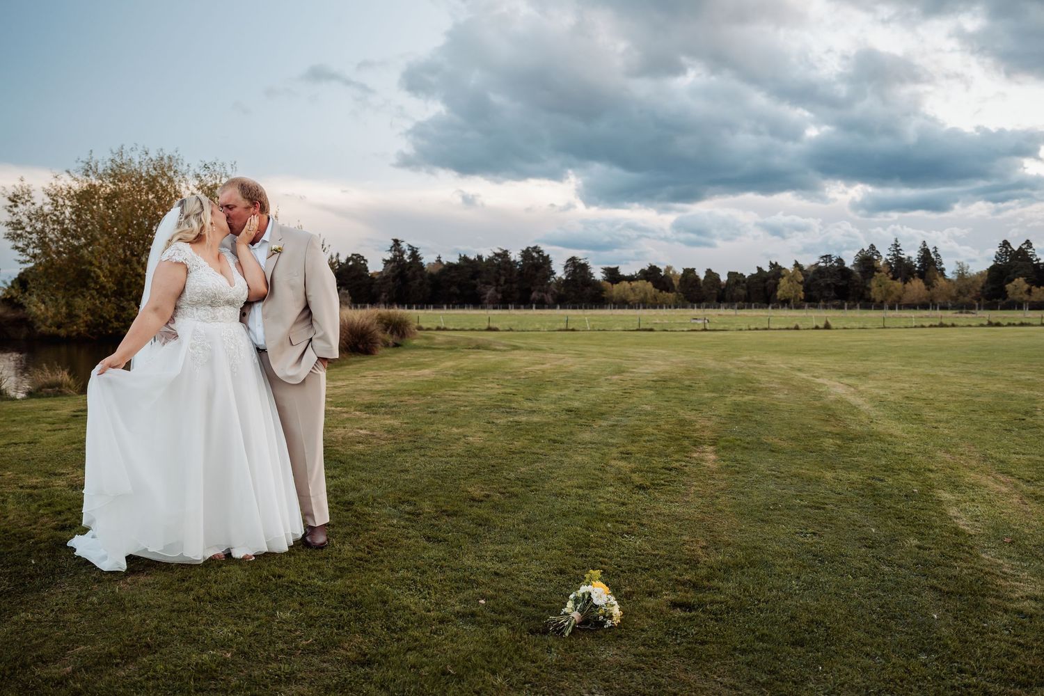 Bride and groom sharing romantic kiss on green lawn under dramatic cloudy sky at outdoor wedding.