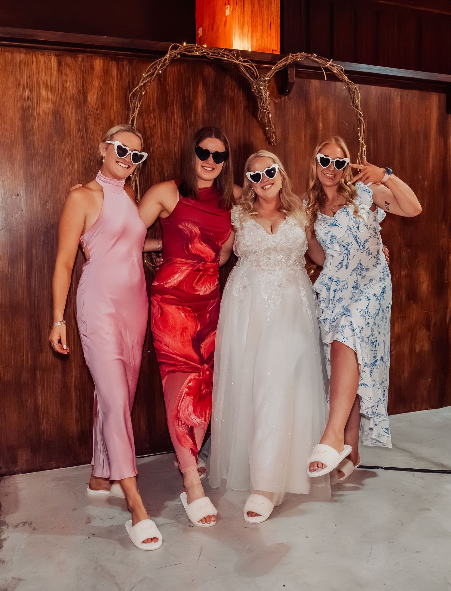Bride in white gown posing with three friends in colorful dresses and heart-shaped sunglasses at reception.