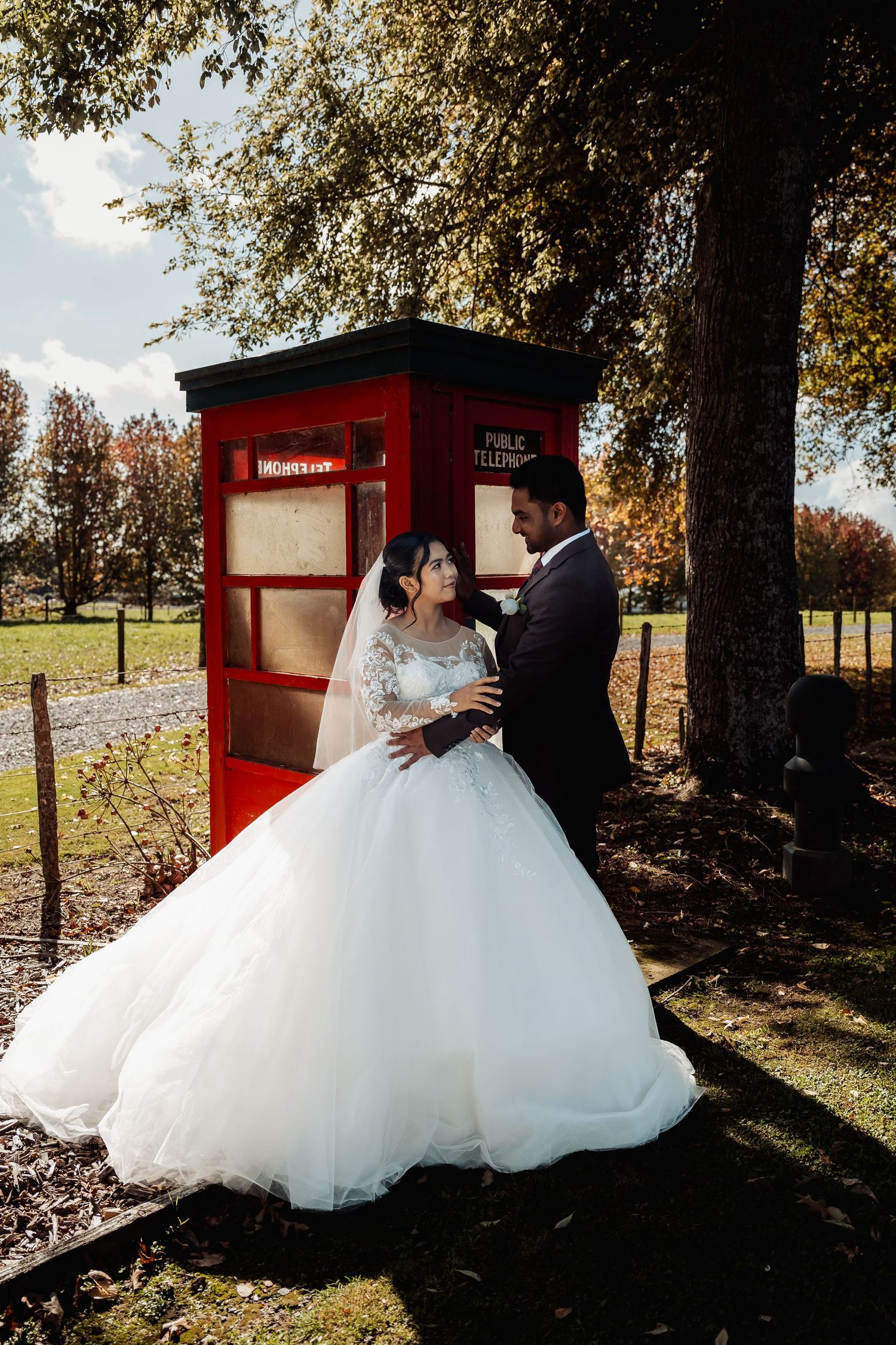 Bride and groom share a romantic moment beside a classic red British phone box in an autumn garden setting.
