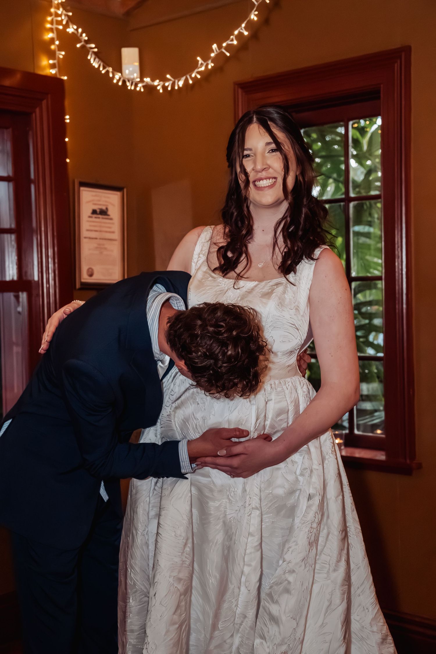 Groom affectionately kissing bride's pregnant belly as she smiles warmly indoors at reception.