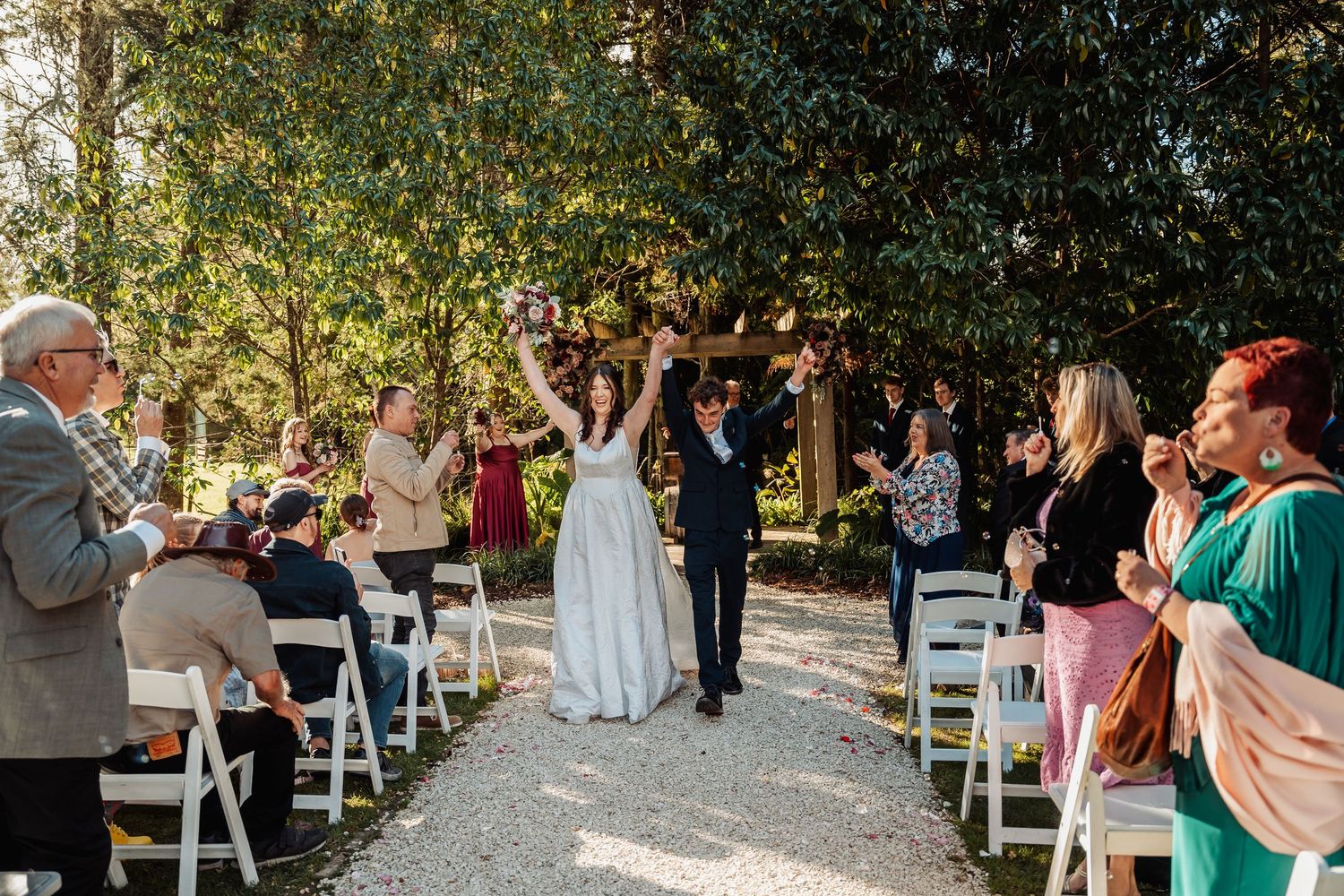 Newlyweds raising hands joyfully while walking down aisle past cheering guests at outdoor wedding.