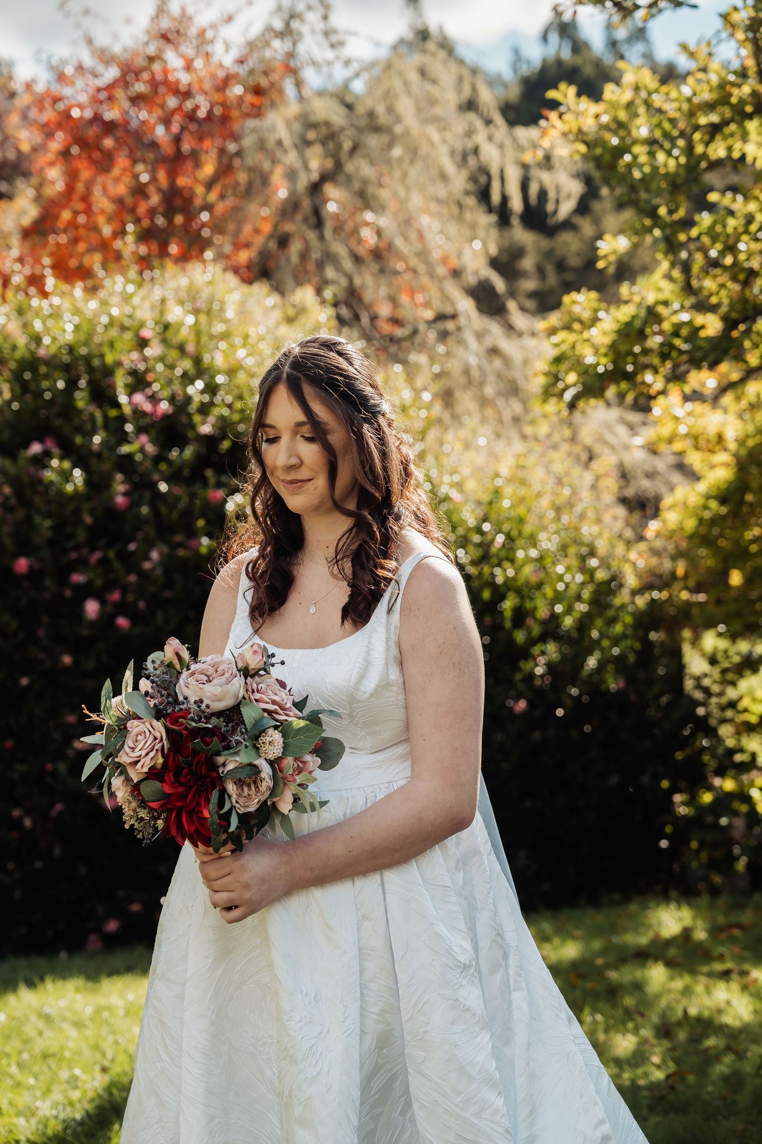Bride in white gown holding bouquet of red and pink flowers in a sunlit garden.