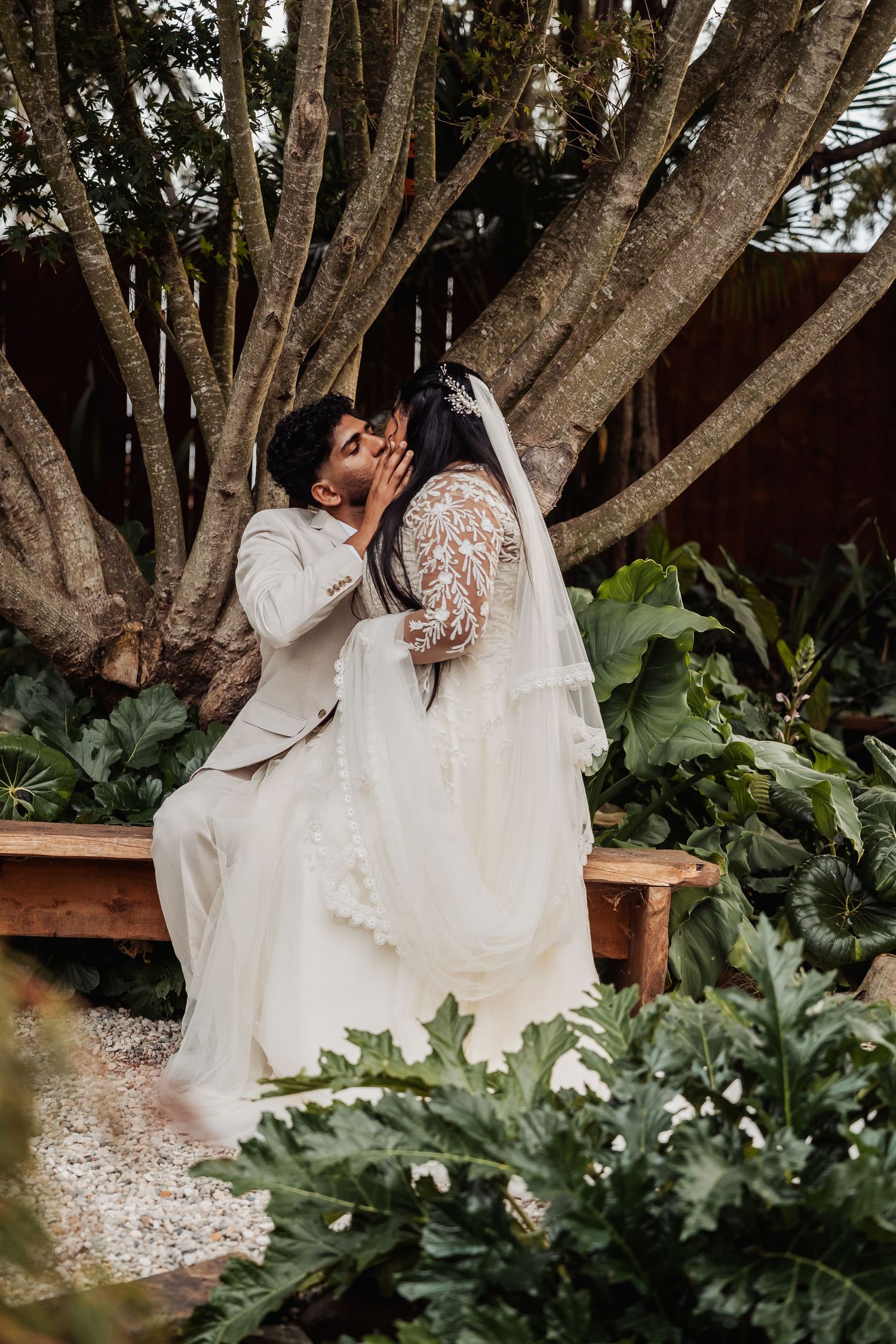 Bride and groom sharing first kiss seated on garden bench surrounded by tropical green foliage.