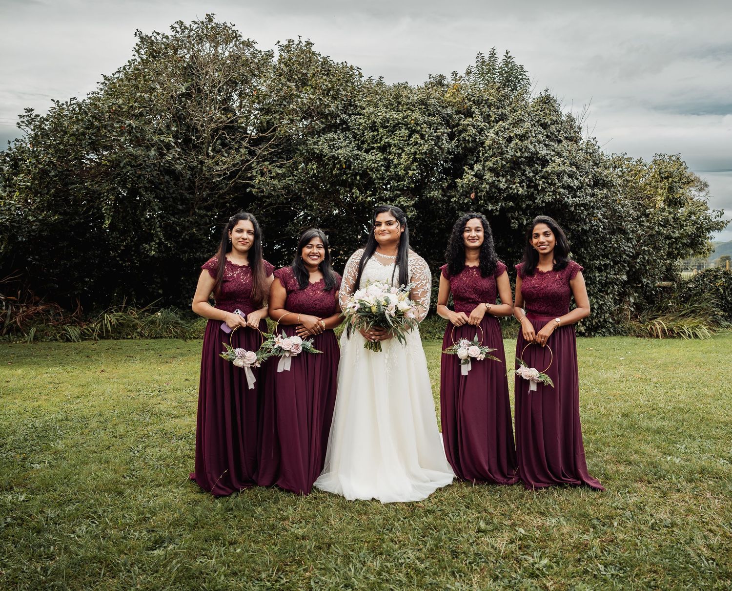 Bride in white gown poses with four bridesmaids in burgundy dresses outdoors on green lawn.