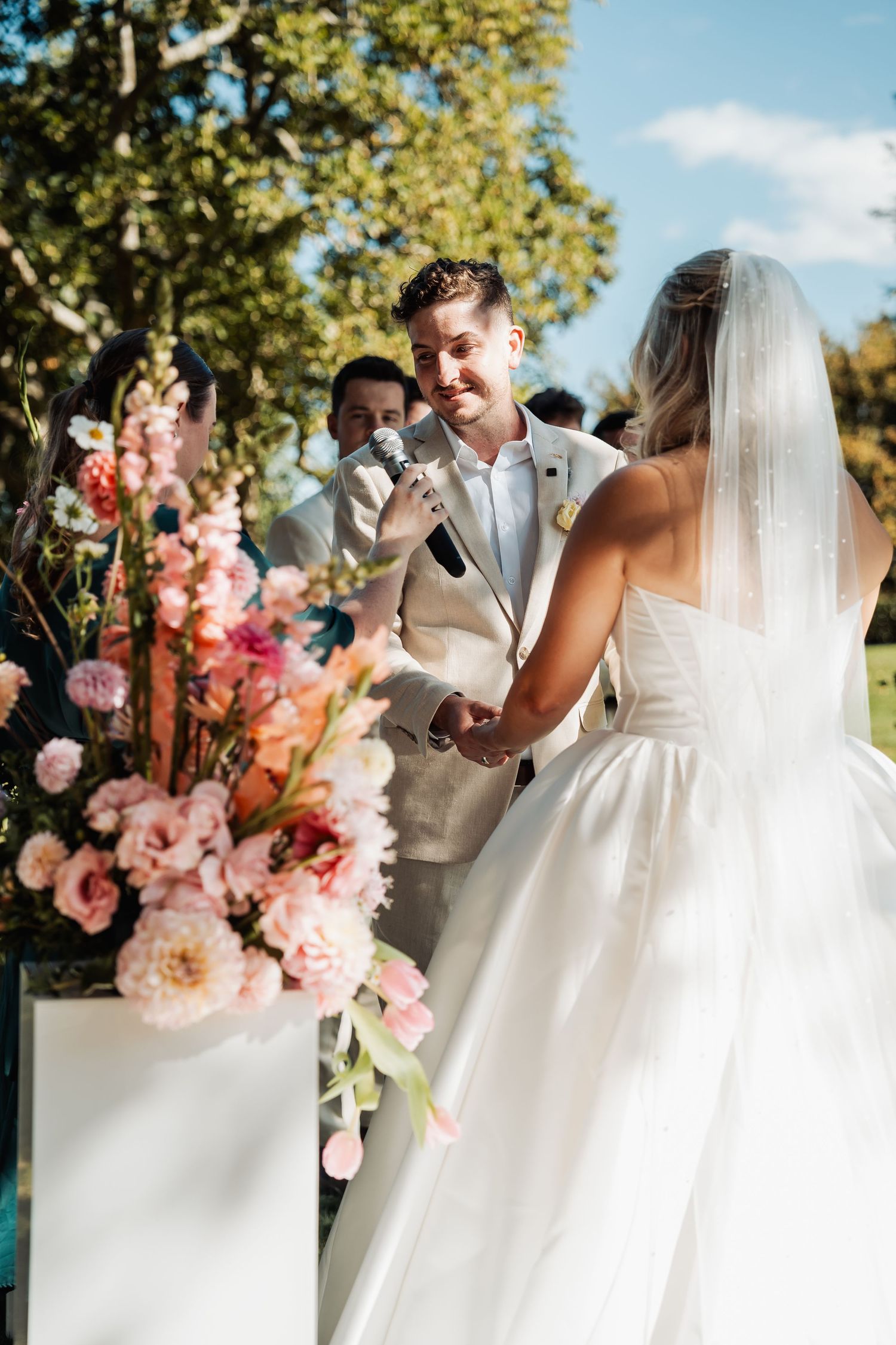 Groom smiles at bride while holding hands during outdoor wedding ceremony with pink floral arrangements.