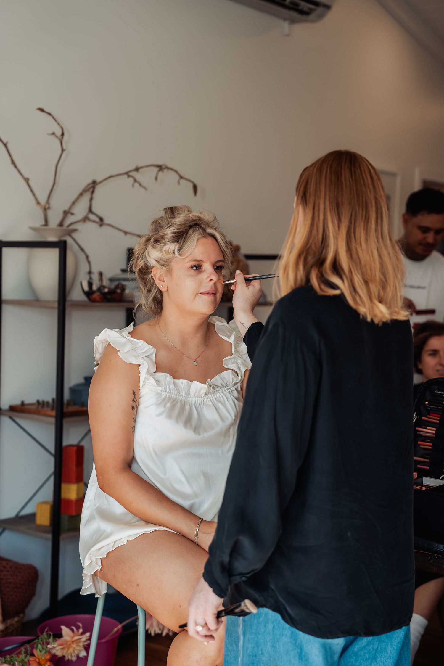 Bride getting makeup applied by a makeup artist in a bright salon before her wedding day.
