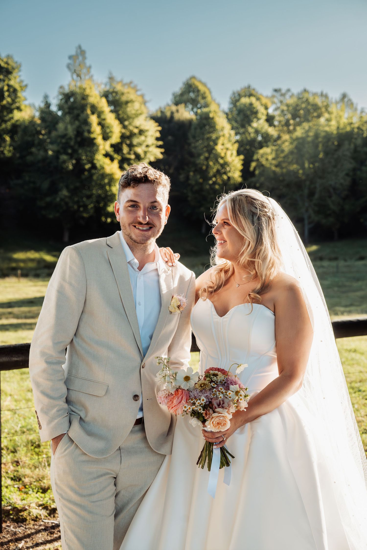 Smiling bride and groom pose outdoors in sunlight, she holds a pastel bouquet, he wears a light beige suit.