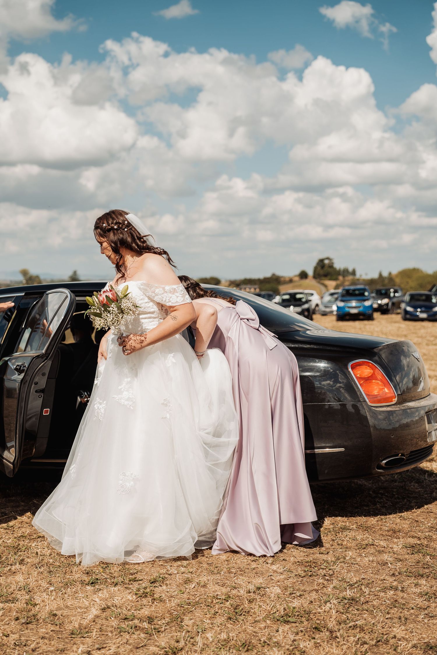 Bride in white off-shoulder gown stepping out of black luxury car assisted by bridesmaid in lavender dress.