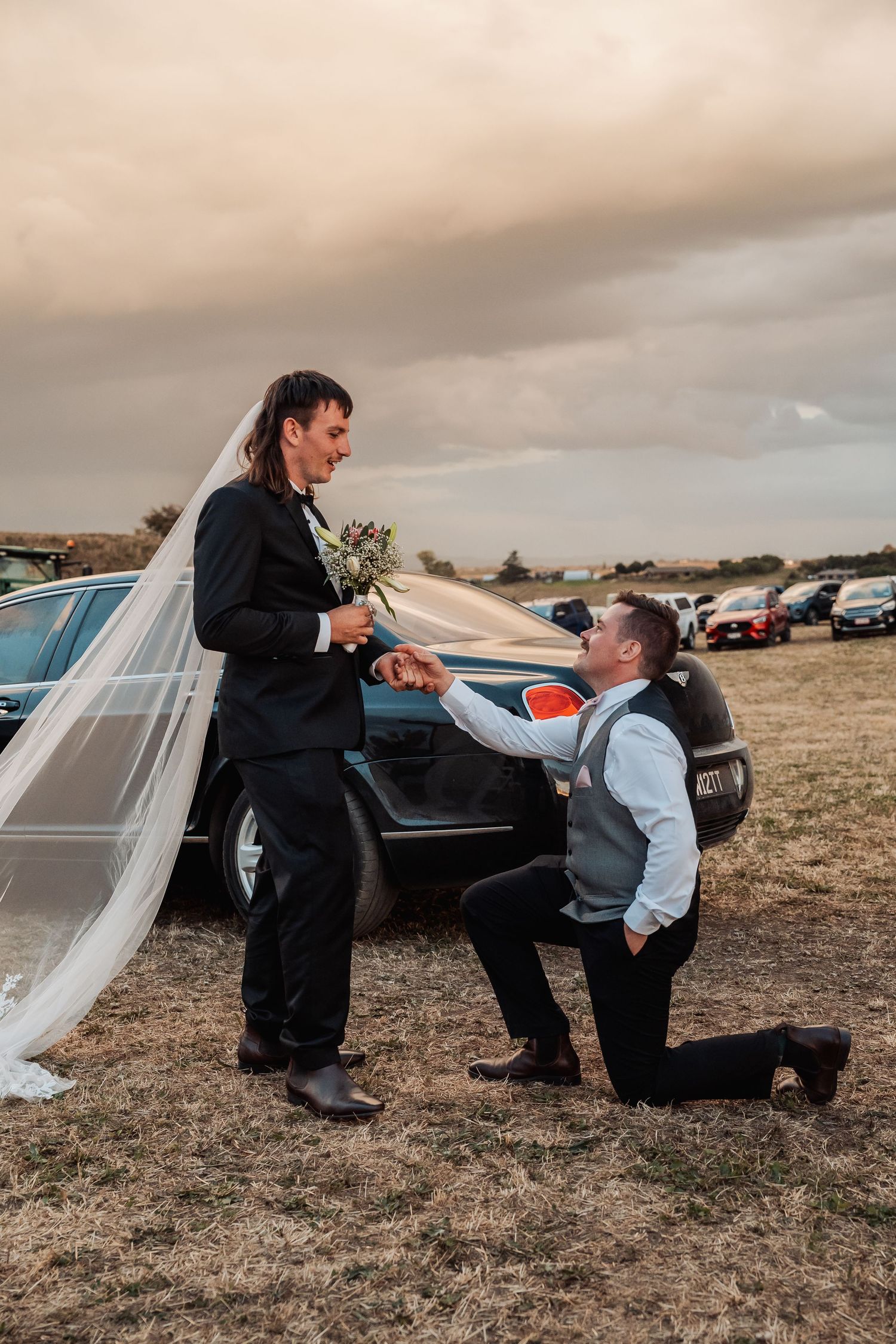 Groomsman kneeling playfully before the groom in a humorous wedding party moment outdoors.