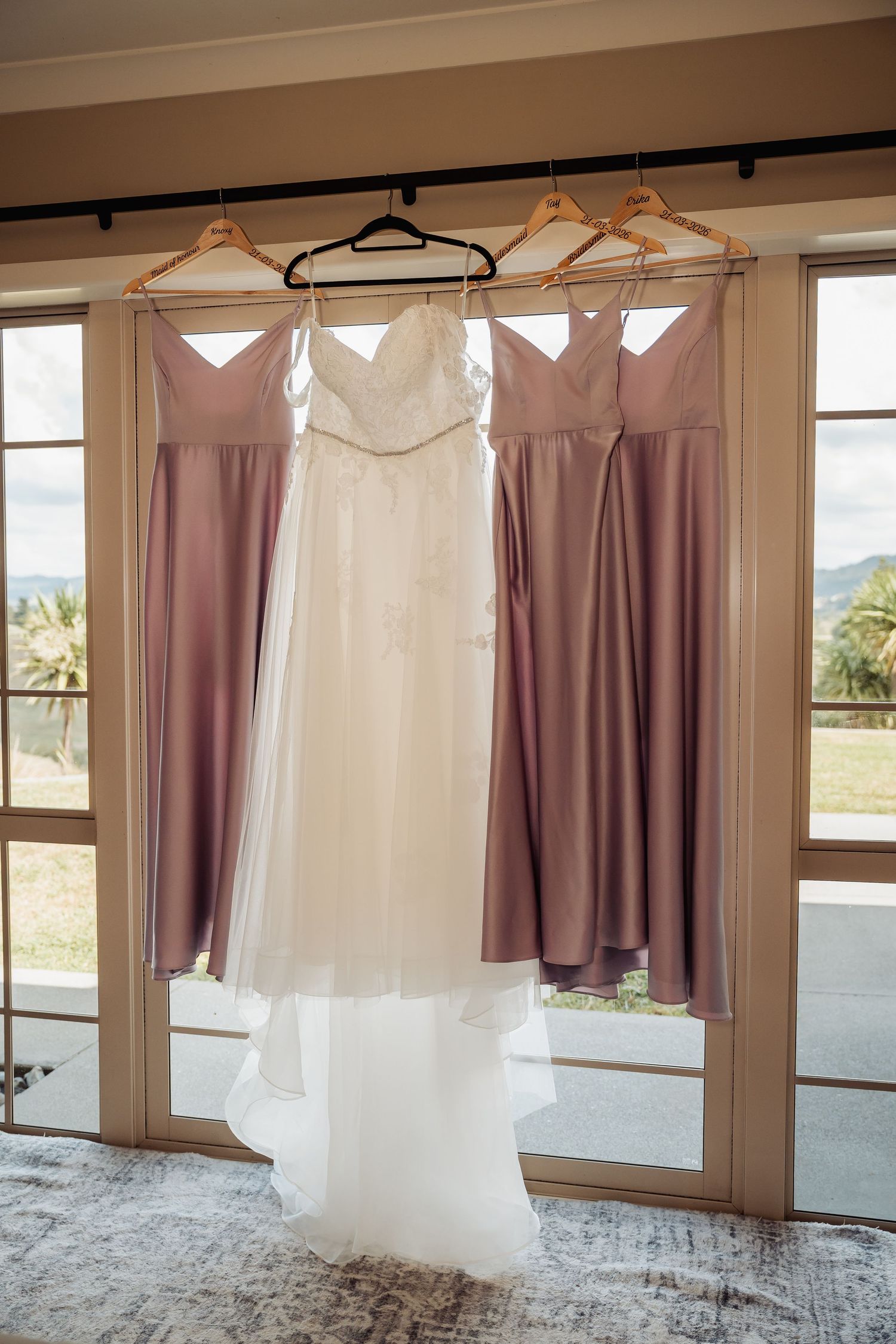 Wedding dress and bridesmaid gowns in dusty rose hanging on wooden hangers before the ceremony.