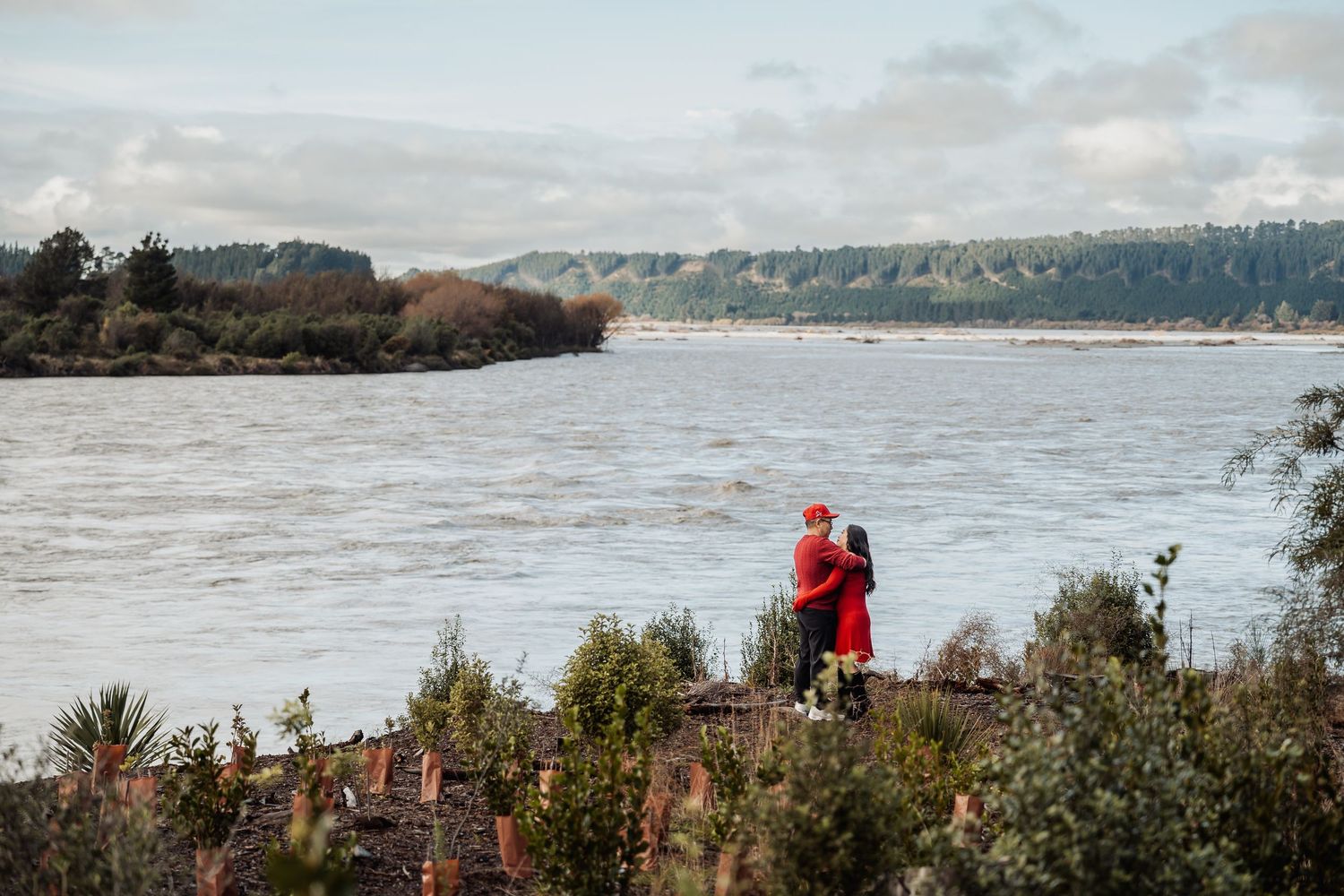Couple embracing on rocky shore overlooking a wide river with lush green trees in the background.