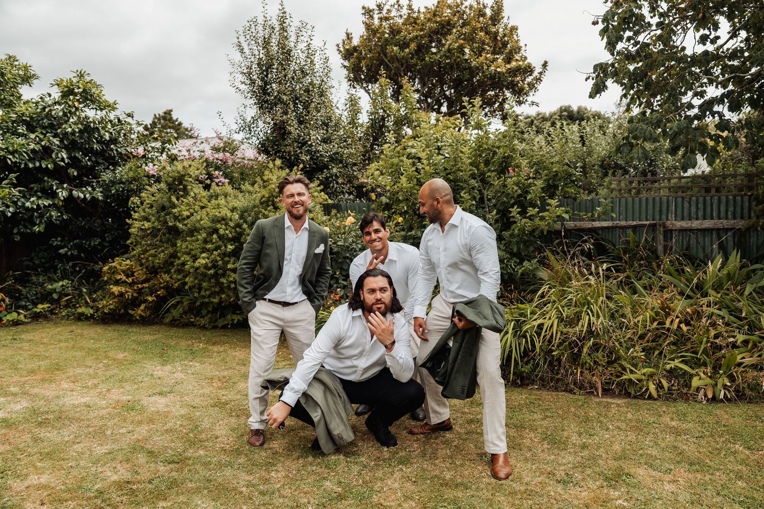 Four groomsmen laugh and pose playfully on a green lawn surrounded by garden foliage.