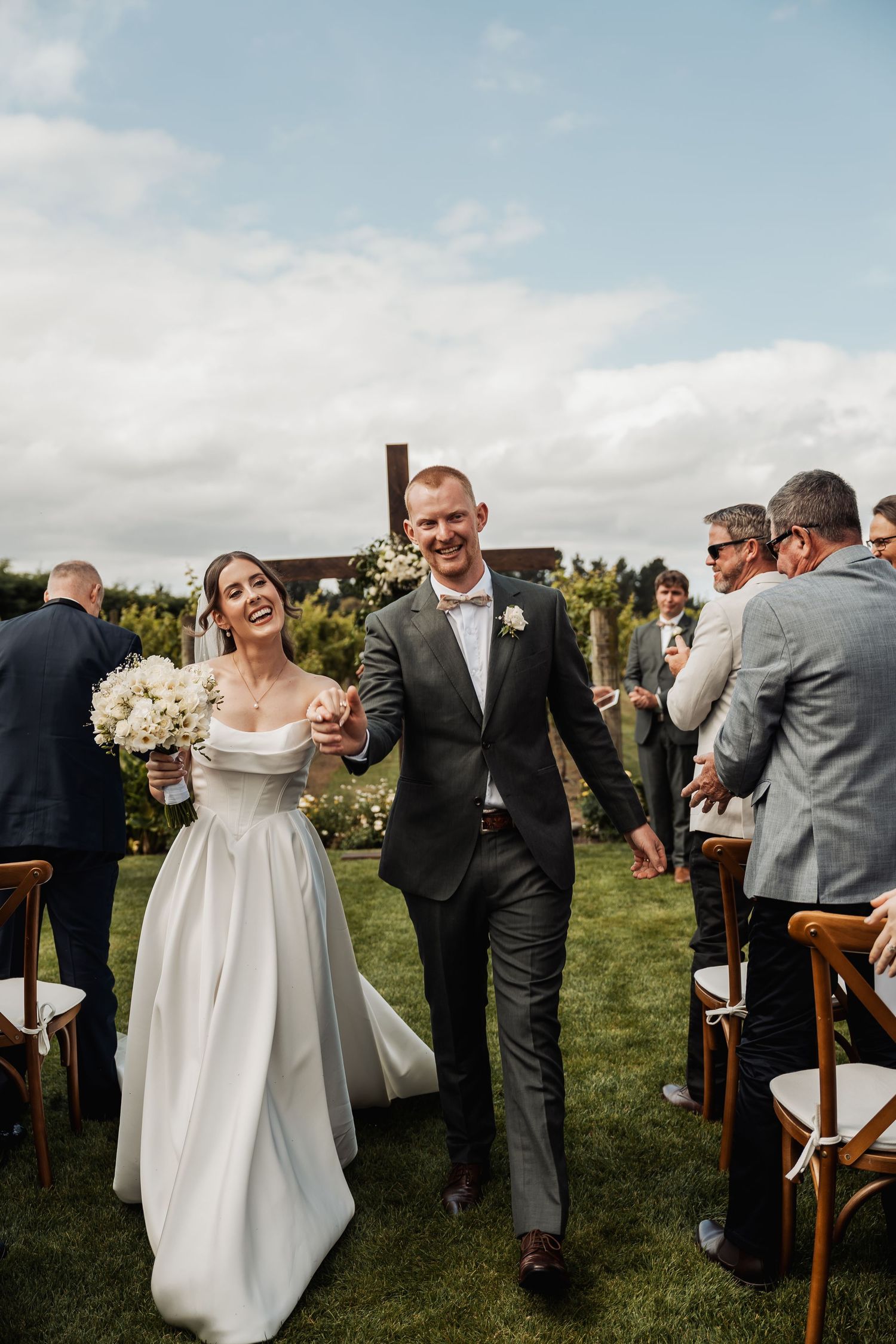 Smiling bride and groom exit outdoor ceremony aisle surrounded by guests at vineyard wedding.