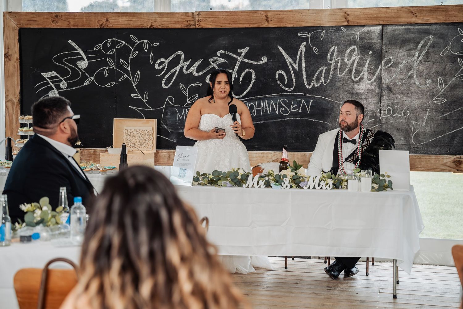 Bride speaking at wedding reception head table beneath Just Married chalkboard sign.