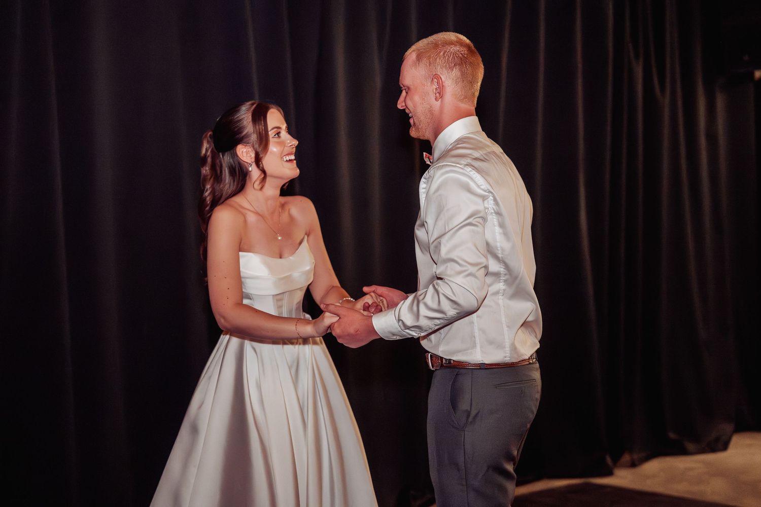 Bride and groom share a joyful first dance moment, holding hands and smiling at each other at their wedding reception.